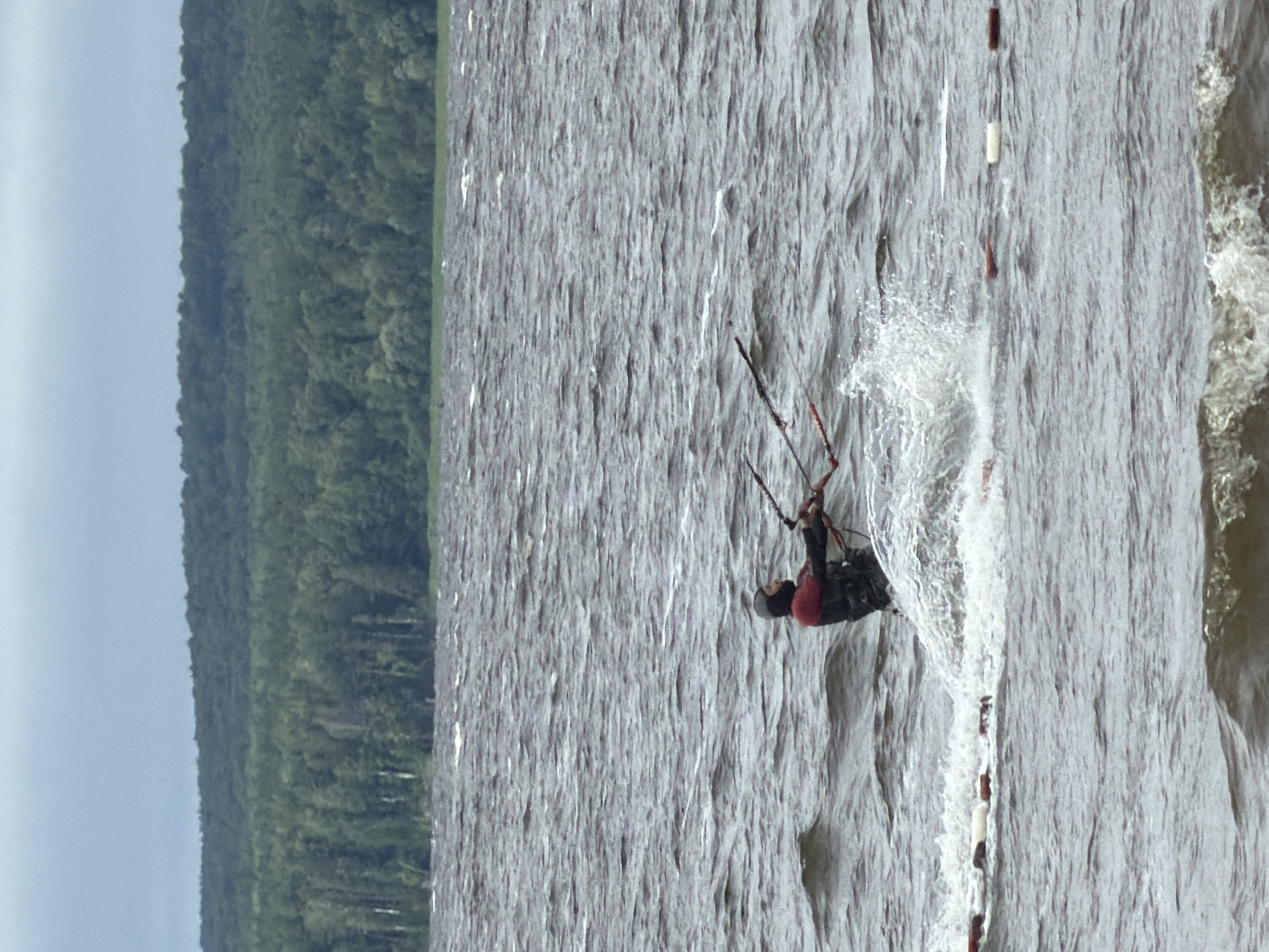 A lone kitesurfer is captured mid-action on a choppy body of water, likely the Kama River in Chaikovsky, Russia. The overcast sky suggests a cool, perhaps breezy, day. The kitesurfer, wearing a red and black wetsuit and a life vest, is actively engaged in the sport, their kite obscured by the frame. Water splashes dramatically around them, indicating a powerful maneuver or a gust of wind. In the background, a dense, dark green forest lines the horizon, meeting a pale, hazy sky. The foreground shows a line of buoys or markers floating on the water's surface, suggesting a designated area or a course. The overall scene conveys a sense of solitary adventure and the raw power of nature.