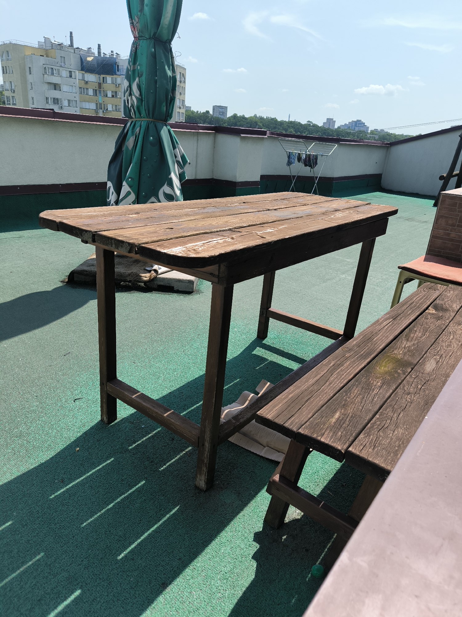 This media file depicts an outdoor scene on a rooftop, likely in Chișinău, Moldova, on a bright, sunny day with a clear blue sky and scattered clouds. The primary subjects are a weathered wooden picnic table and a wooden bench, both appearing rustic and well-used. The table has a slatted top and dark wooden legs, casting long, distinct shadows on the vibrant green, textured rooftop surface. Next to the table, a partially folded, large green umbrella with a white floral pattern is visible, tied with twine.
In the background, a row of buildings with a mix of light-colored facades and some with darker tiled roofs are visible. Some buildings have numerous windows and air conditioning units. Further in the distance, more buildings and a line of green trees suggest an urban or suburban environment. To the right of the table, a portion of a chair with a red seat and light-colored legs is partially visible, and behind it, a brick structure. A clothes drying rack with a few items of clothing is also present in the middle ground, hinting at domestic activity.
The overall impression is one of a relaxed, possibly residential or restaurant rooftop space. The bright sunlight and clear sky indicate daytime, likely midday or early afternoon, given the length and angle of the shadows. There is no visible text in the image. The scene evokes a sense of a warm, pleasant day, with the weathered furniture suggesting a place that has been enjoyed over time.