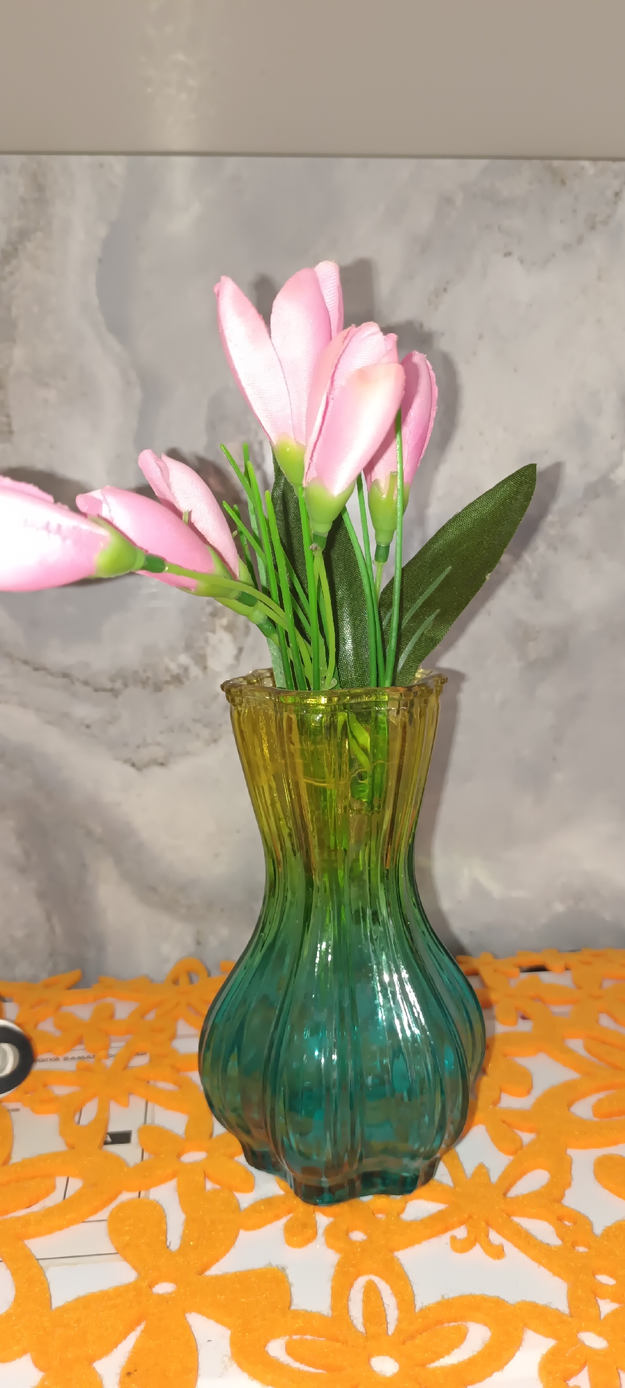A close-up, vertical shot shows a glass vase with pink artificial flowers and green stems and leaves. The vase has a ribbed texture and a gradient color from yellow at the top to green at the bottom. It sits on an orange, floral-patterned placemat. The background is a gray, textured wall. The lighting is soft, and there are no people or animals visible. The scene appears to be indoors, likely in a domestic setting, possibly in Fergana City, Uzbekistan.