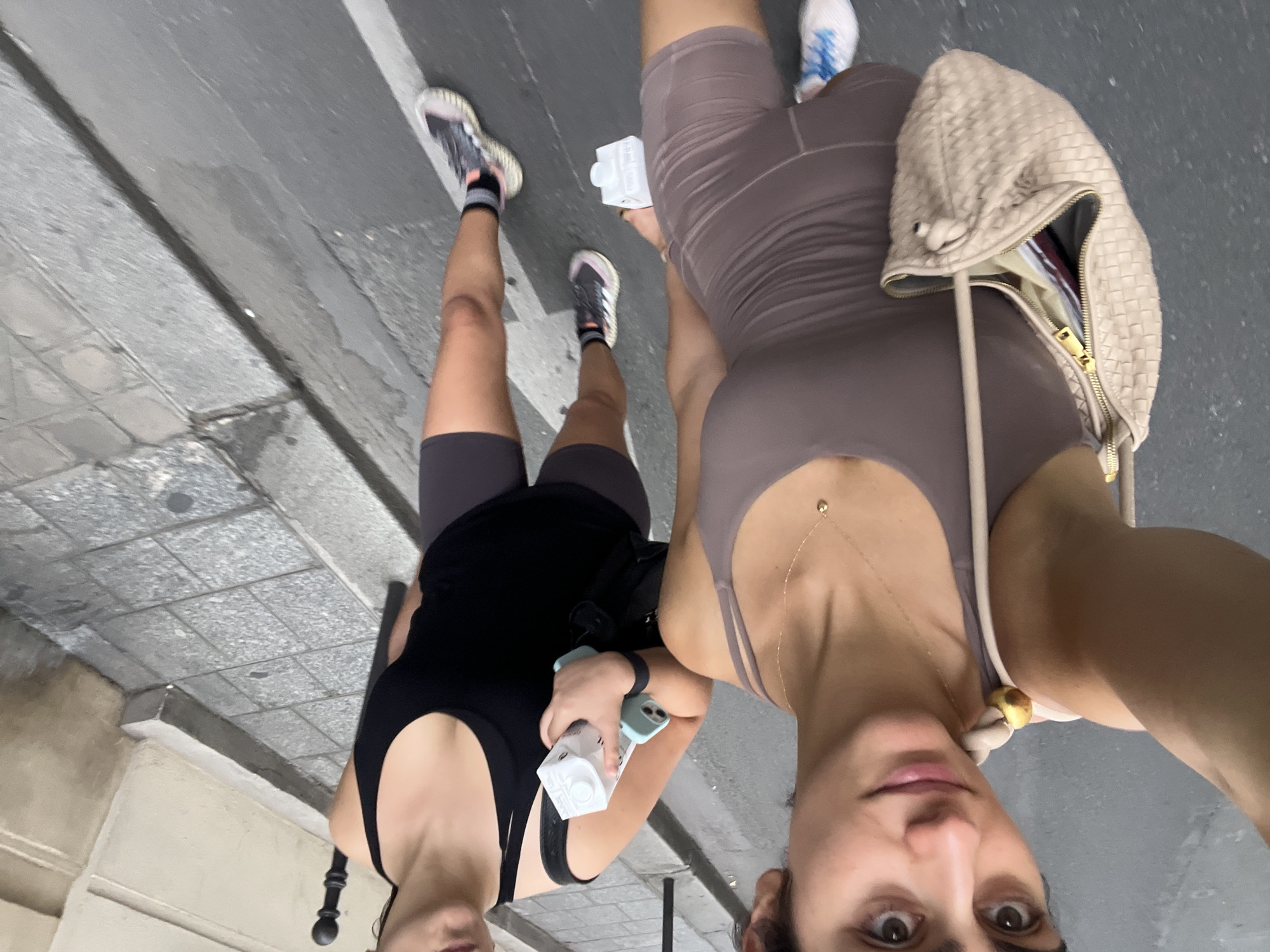 Two people are walking side-by-side on a street in Paris, France. The person on the left is wearing a black tank top and dark grey cycling shorts. They have dark hair pulled back and are holding a white carton of liquid and a light blue phone. The person on the right is wearing a mauve tank top and matching cycling shorts. They have dark hair and are wearing a delicate gold necklace. They are carrying a light beige woven handbag over their shoulder. Both individuals are wearing athletic shoes and appear to be in motion, suggesting they are walking or perhaps jogging.
The scene is an urban street with a grey asphalt road and a textured grey tiled sidewalk. A white line is painted on the road. The lighting suggests it is daytime, possibly overcast or late afternoon given the soft light. There is no visible text in the image. The overall mood is casual and active, consistent with people out for a walk or exercise in a city.