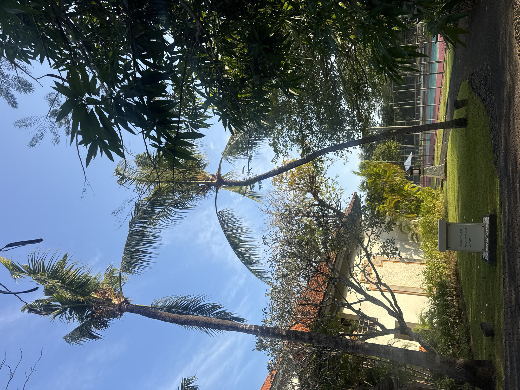 The image captures a serene outdoor scene in Kuta Selatan, Indonesia, likely within a resort complex. Tall palm trees and lush green foliage dominate the foreground and midground, with a clear blue sky peeking through. A building with a terracotta-tiled roof is visible on the left, partially obscured by trees. In the midground, a tennis court with a red and green surface and a chain-link fence is present. A small, white directional signpost with text indicating "WEST VILLAGE," "SOUTH VILLAGE," and "LOBBY" is situated on a manicured lawn. The overall atmosphere is tranquil, suggesting a tropical vacation setting. There are no people or animals visible in the image.