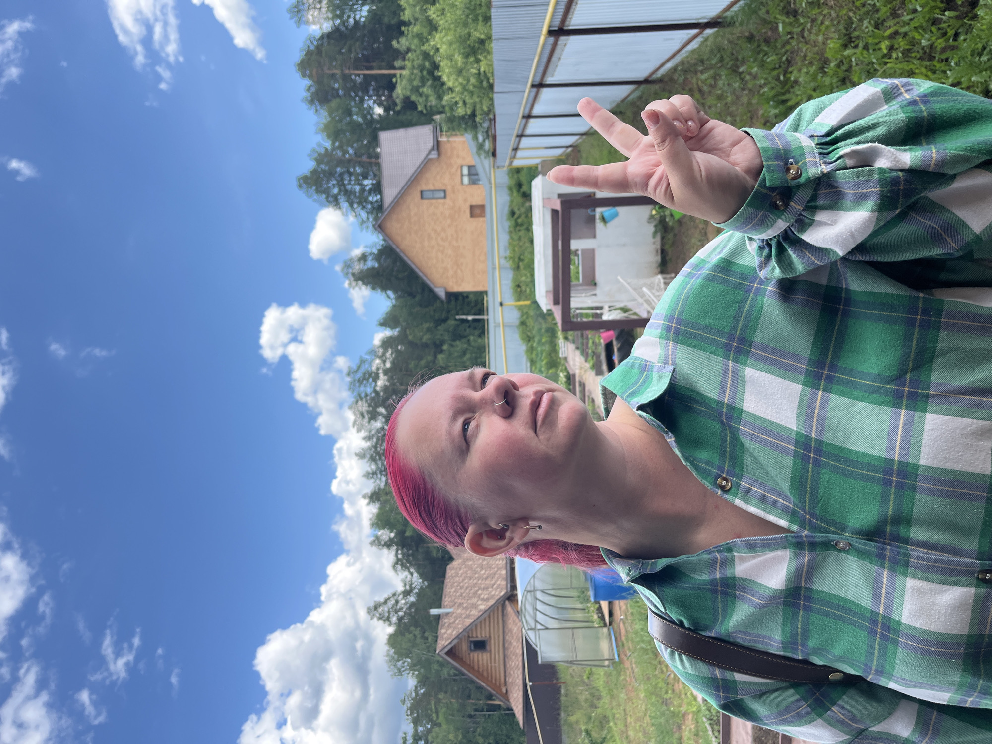 An adult female, positioned slightly off-center to the left, is captured outdoors in the city of Chaikovsky, Russia. She possesses vibrant pink hair, a nose ring on her left nostril, and multiple piercings in her left ear. She is wearing a long-sleeved, collared shirt featuring a green, white, and blue plaid pattern with gold buttons; the sleeves are puffed at the cuffs. A brown strap is visible over her right shoulder. Her right hand is raised, with the index and middle fingers extended to form a peace sign. Her gaze is directed towards the right side of the frame, slightly upwards.
The background consists of a bright blue sky with scattered white cumulus clouds. Below the sky, dense green trees line the horizon. On the left, a two-story house with light brown wooden siding and a dark roof is visible. To its right, a tunnel-shaped, transparent greenhouse with an open door and a blue barrel nearby is situated in a grassy area. Further to the right, a corrugated metal fence with a yellow top rail extends across the midground. Behind this fence and further into the distance, another two-story house with light orange/brick siding and a dark roof is discernible. Green grass and various low-lying plants cover the foreground and midground. A small, white, cuboid structure with a brown open frame on top is visible to the right of the subject, near the fence.