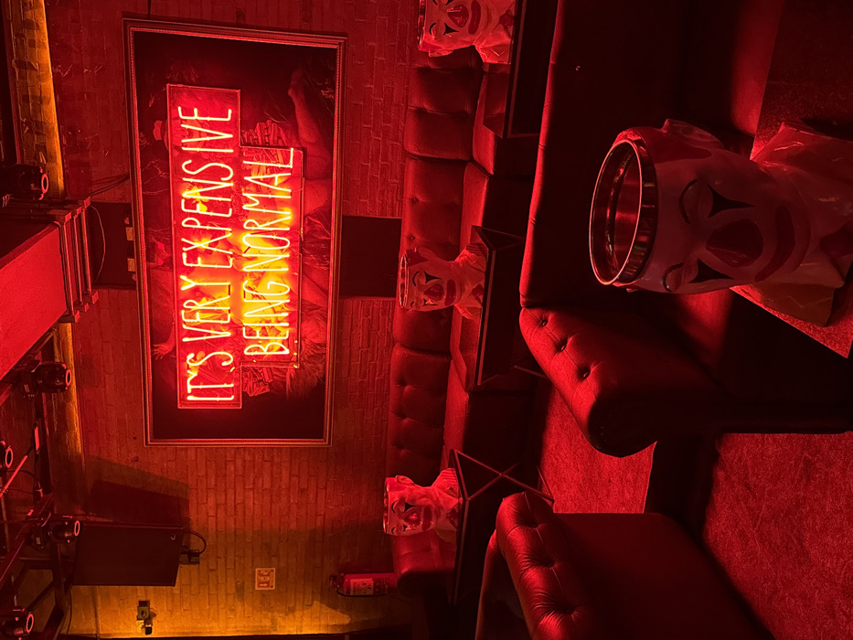 A dimly lit, rotated image showcases a lounge area bathed in red light. A neon sign reads, "It's very expensive being normal." Several clown mask sculptures are positioned along a dark tufted couch. A metal-rimmed container sits on the couch beside a mask. Visible equipment and brick walls suggest an industrial-chic aesthetic. The overall atmosphere is eerie.