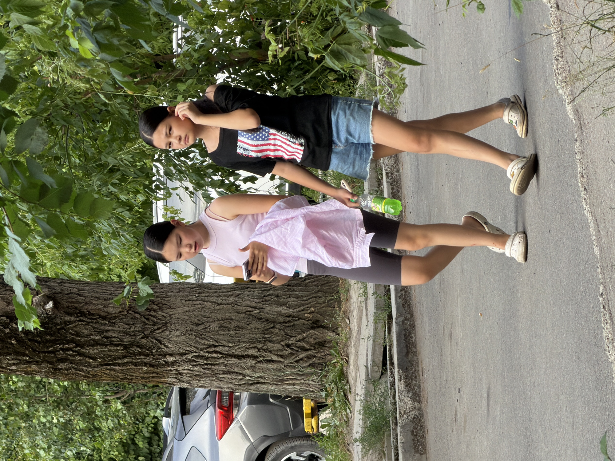 Two young girls are walking on an asphalt path next to a tree and some vegetation. The girl on the left is wearing a pink sleeveless top, grey shorts, and white clogs. She is holding a pink cloth and looking at her phone. The girl on the right is wearing a black off-the-shoulder shirt with an American flag design, denim shorts, and white clogs. She is holding a green bottle of drink and touching her ear. A grey SUV is parked behind them. The environment is an outdoor urban setting.