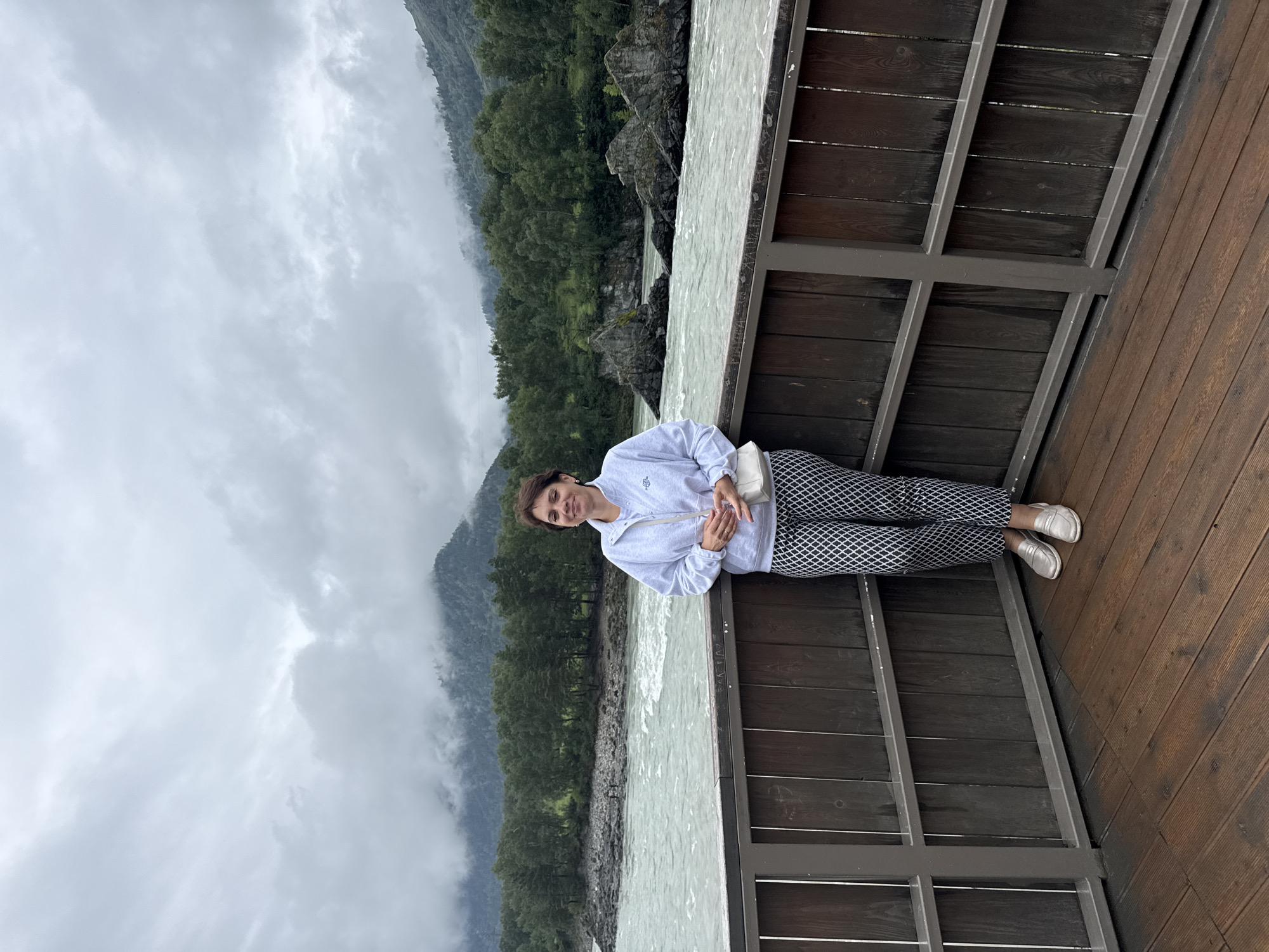A woman stands on a wooden deck in front of a railing, with a river and mountainous landscape in the background. She is wearing a light gray hooded sweatshirt, patterned black and white pants, and silver slip-on shoes. She has a white purse slung over her shoulder. The river is wide and appears to be flowing rapidly, with rocky outcrops visible in the water. The mountains are covered in dense green forest and are partially obscured by clouds. The sky is overcast. The location is identified as Elekmonar, Russia.
