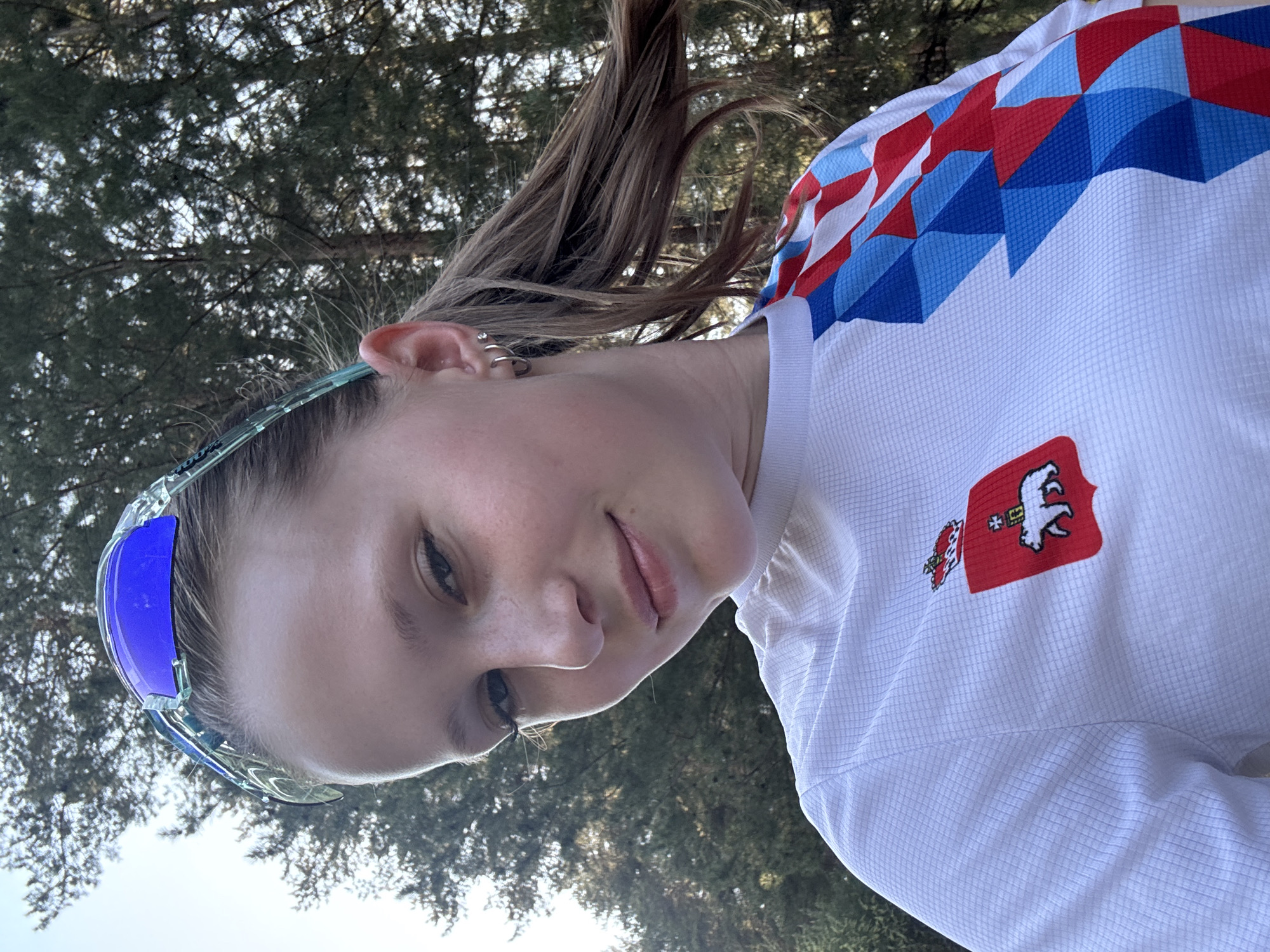 A young woman is posing for a selfie outdoors in a sunny location with trees in the background. She is wearing cycling glasses with blue lenses and a white cycling jersey with a red and blue geometric pattern on the shoulders and a red patch featuring a white bear on the chest. The patch likely represents the city of Perm Krai, Russia, of which Chaikovsky is a part. The location seems to be a park or wooded area.