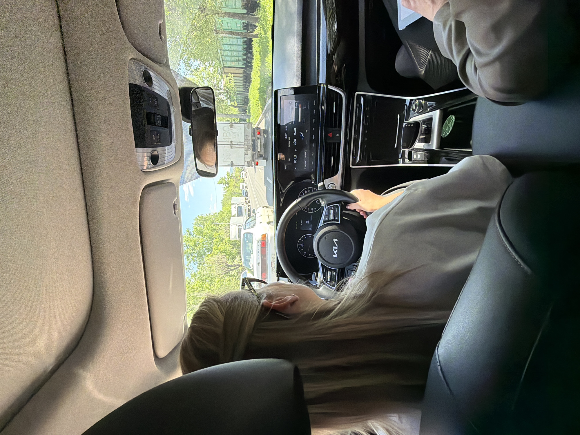 The image shows the interior of a car, specifically the driver's seat and dashboard area. A blonde-haired woman with glasses is driving the car. She has her hands on the steering wheel, which features a Kia logo. The dashboard includes a digital display screen. Through the windshield, a view of a road with other vehicles is visible, including trucks. A train wagon can be seen in the background. The location is Krasnoyarsk, Russia.