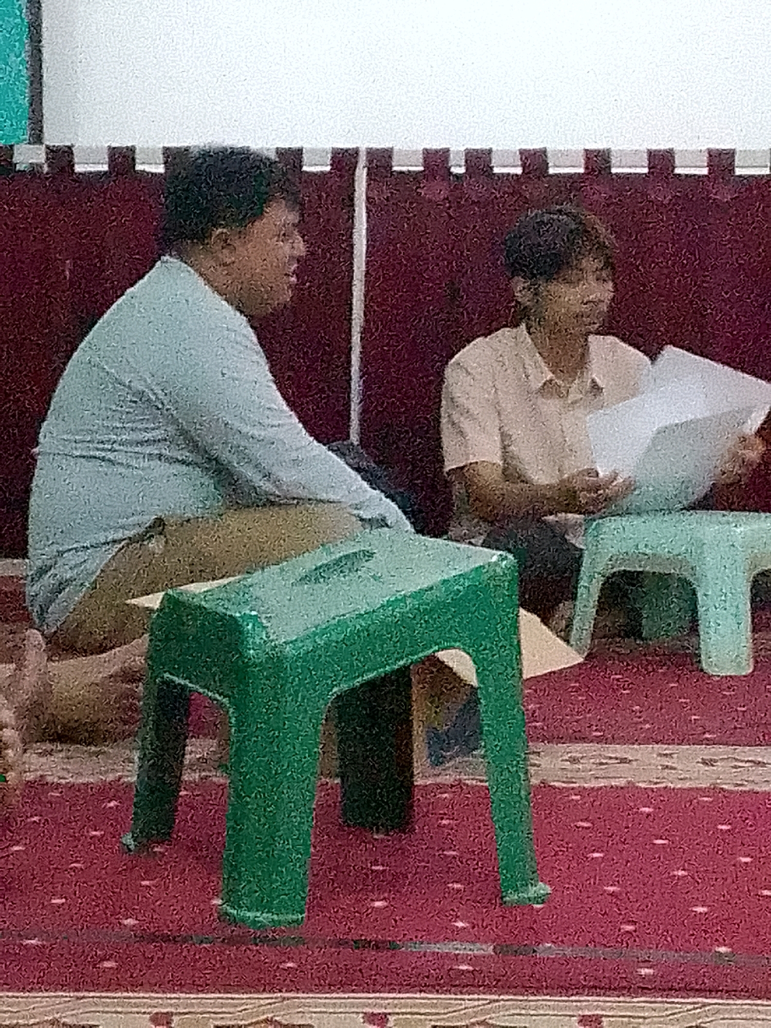The image depicts two individuals indoors, seated on a red carpet. In the foreground, a person with dark, curly hair, wearing a light blue long-sleeved shirt and tan pants, is sitting with their legs crossed. To their right, another individual, with shorter, dark hair and wearing a light pink collared shirt, is seated and holding white papers, appearing to be reading or discussing them. Between the two individuals are two small, brightly colored plastic stools, one green and one light blue. The background features a dark red wall with a decorative pattern of white and black squares. The lighting appears to be overhead, casting a shadow from the green stool onto the carpet. The overall scene suggests a casual meeting or discussion taking place in an indoor setting.