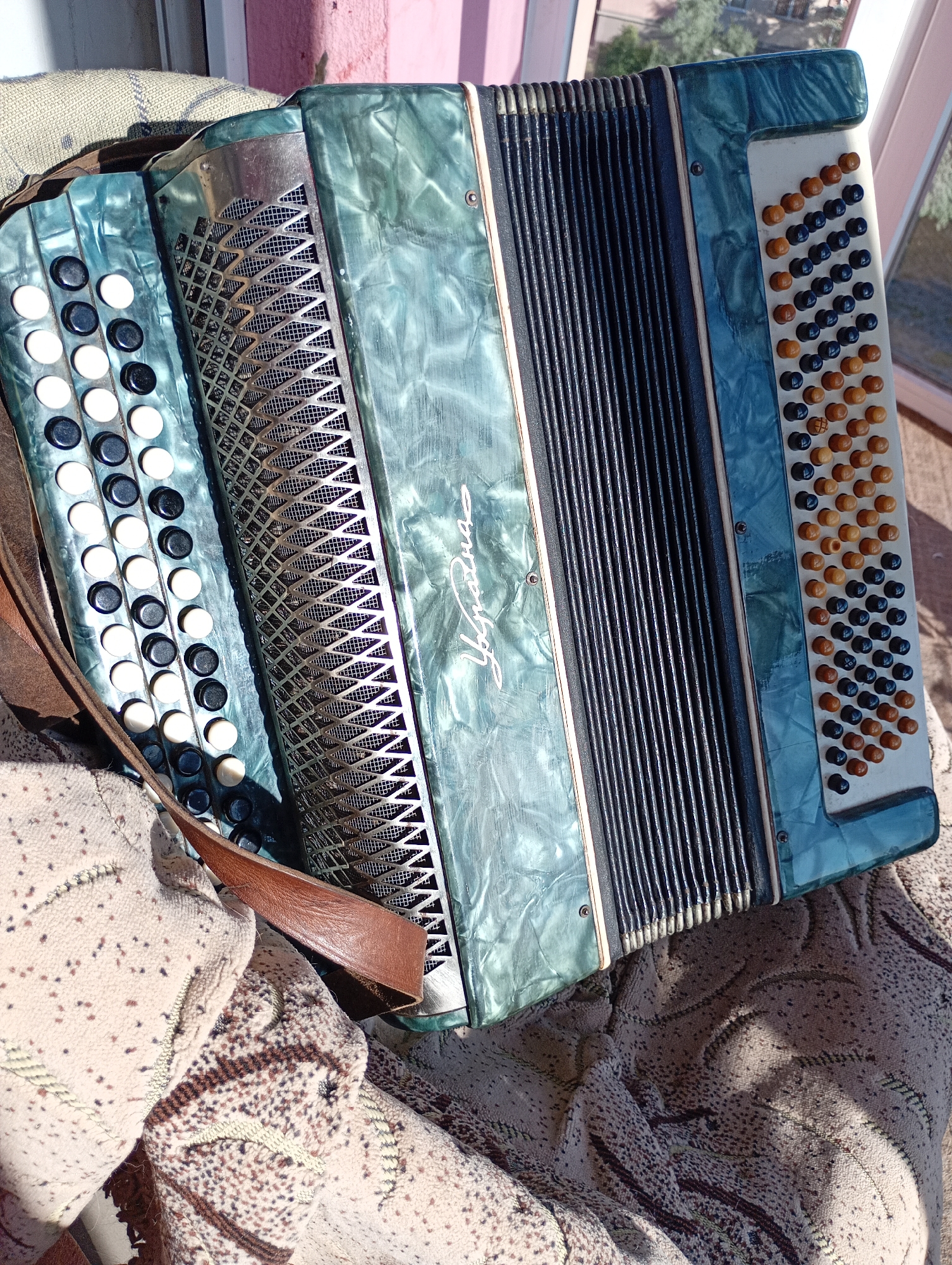 An accordion is placed on a patterned fabric surface, likely outdoors, with sunlight casting shadows and highlighting the instrument. The accordion is a turquoise color with black and white keys on the left side and orange and black buttons on the right. A brown leather strap is attached to the instrument. In the background, a pink wall and a window with trees are visible, suggesting an urban or suburban setting within the city of Avanhard, Ukraine. The image is a close-up, focusing on the accordion and the fabric.