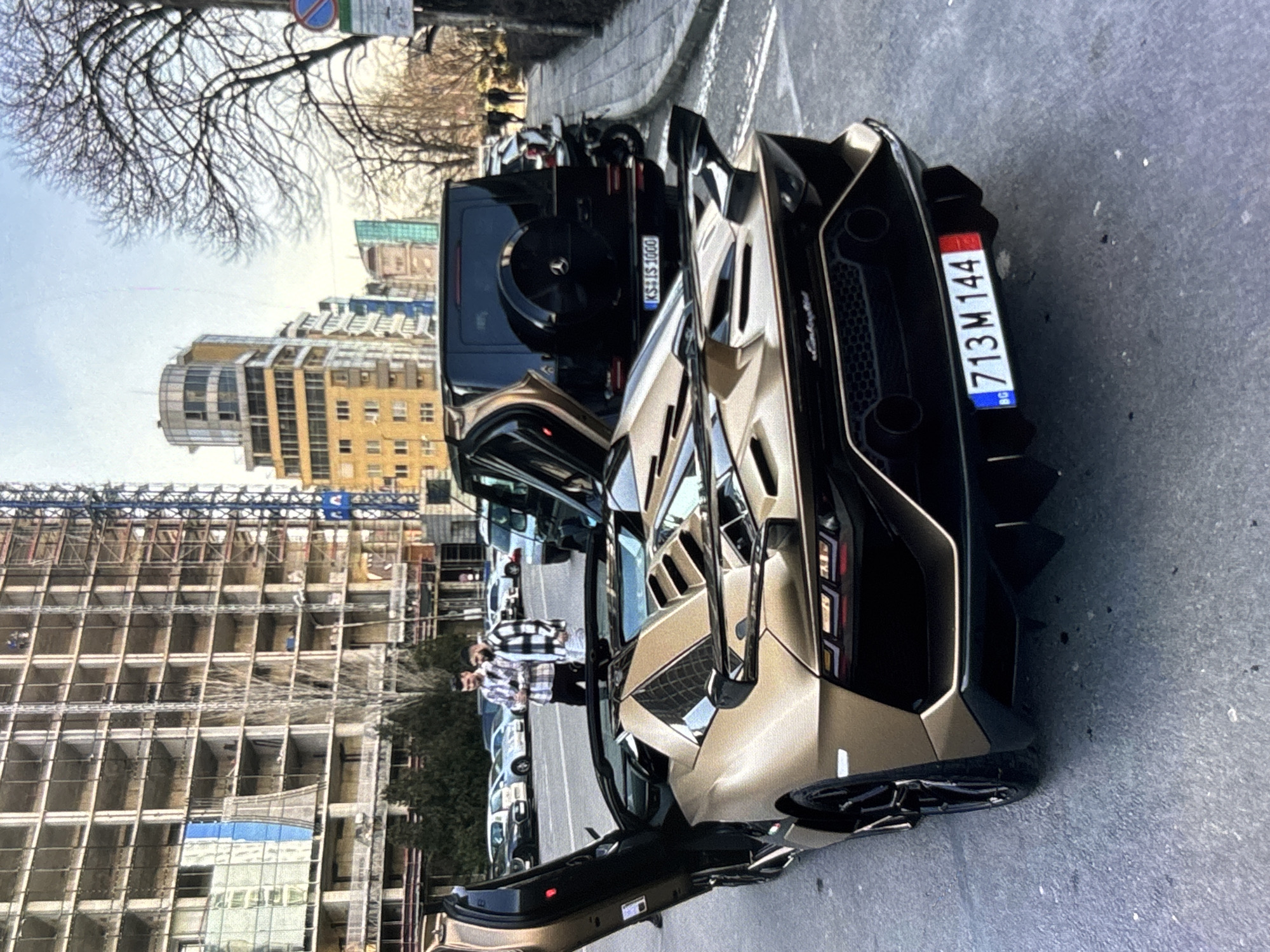 This urban street scene prominently features two luxury SUVs parked on an asphalt road during daylight hours. In the foreground, a gold/bronze Lamborghini (likely an Urus) with its driver's side door open, reveals a glimpse of its interior. The car's rear displays a Bulgarian license plate, "713 M 144," with "BG" on the blue strip, and a subtle "Lamborghini" script. Behind it, a black Mercedes-Benz G-Wagon is parked, featuring a German license plate "KS S 1000."
Two people are visible near the open door of the Lamborghini. One person, wearing a black and white plaid shirt, faces away from the camera, seemingly observing the vehicle or their surroundings.
The background showcases a cityscape with a mix of modern architecture. To the left, a large multi-story building is under construction, exposing its structural framework and scaffolding. Further back, several finished buildings with various designs, including one with a distinctive curved glass section, line the street. Bare trees, suggesting a late autumn or winter setting, are scattered throughout the scene. A blue circular street sign is partially visible against the bright, possibly overcast sky. The overall atmosphere is that of a typical, active city environment.