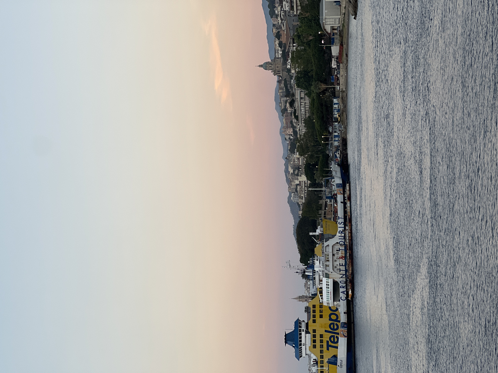 The image captures a panoramic view of the harbor and cityscape of Messina, Italy, at dusk. The sky exhibits a gradient from light blue at the top, transitioning to soft peach and pink tones towards the horizon. The foreground consists of dark, subtly rippled water of the harbor.
On the left side of the frame, two large ferries are docked. The most prominent is a yellow and blue ferry, featuring "Telepass" written in large blue letters on its side and "ZANCLE" near its bow. This vessel has a distinct blue upper deck and bridge structure. Adjacent and partially obscured is a white ferry with "CARONTE & TOURIST" clearly visible on its side. Other smaller vessels are docked nearby.
The mid-ground reveals a densely built-up urban area extending from the waterfront up a series of hills. A dominant feature is a large, light-colored church or sanctuary topped with a prominent green dome, situated on a central elevated position. Further to the left, a tall, slender church spire can be seen rising above the buildings. The cityscape comprises various residential and commercial structures of different heights and architectural styles, interspersed with numerous dark green trees and patches of vegetation. In the background, dark blue silhouettes of distant mountains rise against the horizon.
The immediate shoreline on the right side of the image consists of a rocky or concrete edge, featuring several small, light-colored modular buildings or containers. No human figures are discernible within the scene.