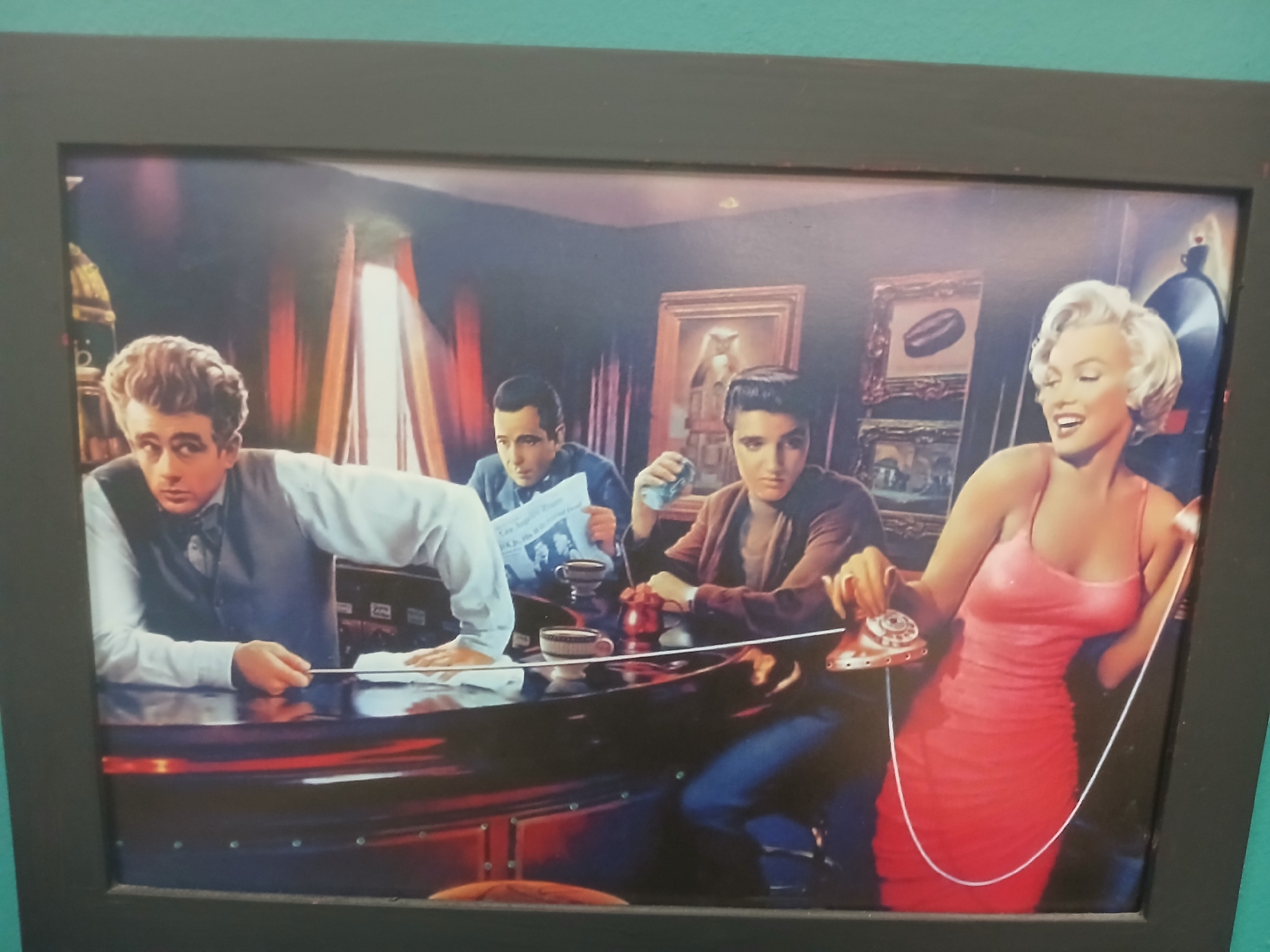 The image depicts a framed, printed artwork featuring four iconic mid-20th-century American celebrities. On the left is a man with curly dark hair, wearing a white shirt and a dark vest, leaning on a dark, reflective surface. To his right is another man in a dark blue shirt, holding a newspaper and a cup. Next to him is a third man with dark hair, wearing a brown jacket, holding a red object. On the far right is a woman with blonde hair, wearing a pink sleeveless dress, holding an old-fashioned rotary telephone. The setting appears to be an indoor space, possibly a bar or lounge, with dimly lit walls, curtains in the background, and framed artwork on the walls. The overall impression is that of a stylized or fictionalized gathering of these personalities. The image is presented as a photograph of the artwork, with a dark frame and a teal wall visible in the background. There is no information about the city or specific location.