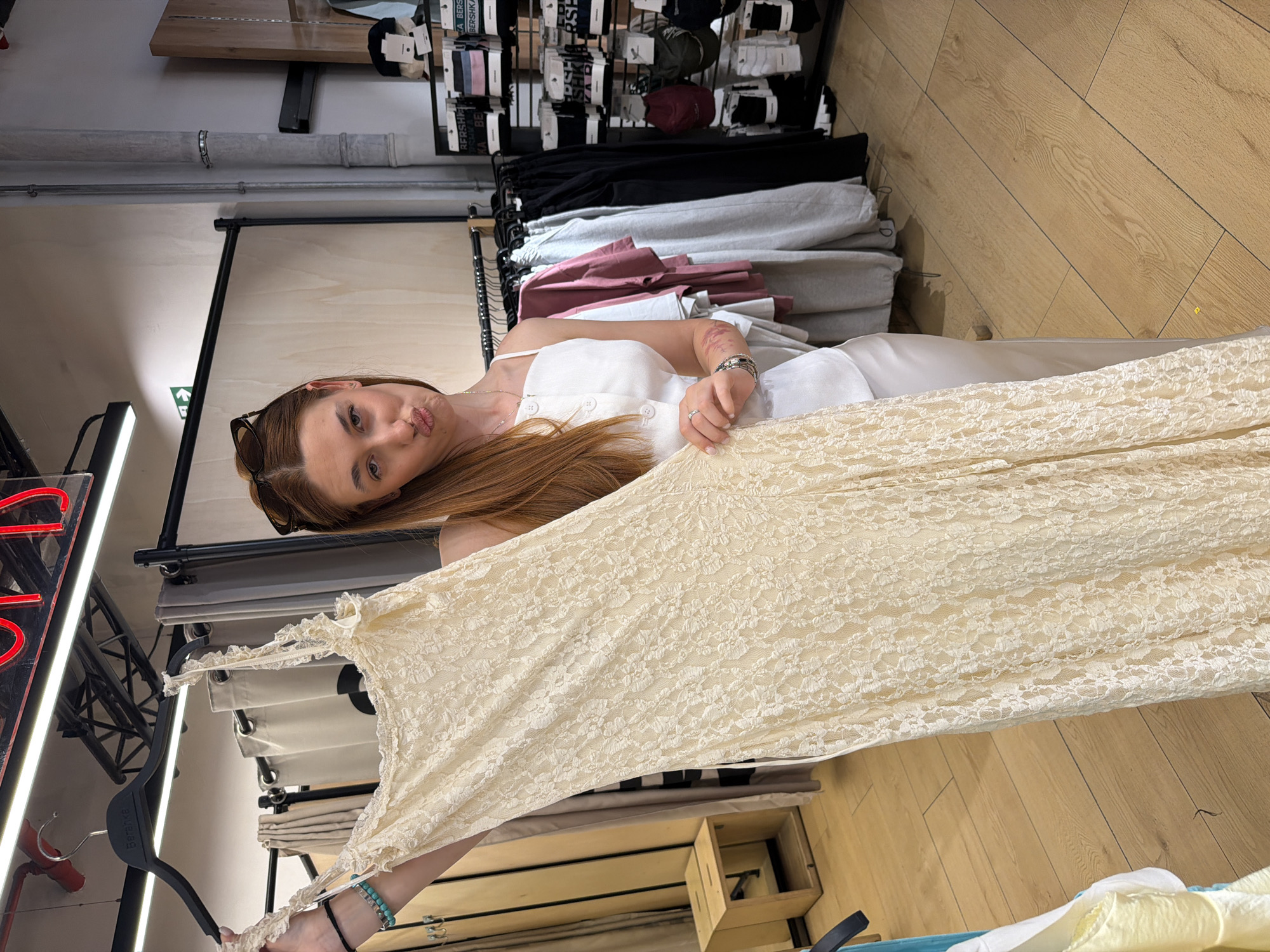 A young woman with long auburn hair and sunglasses on her head is standing in a clothing store in Sofia, Bulgaria. She is wearing a white dress with spaghetti straps and holding up a long, off-white lace dress on a black hanger. The background includes clothing racks filled with various items, including shirts, pants, and accessories. The store has wooden floors and overhead lighting. A red neon sign is visible in the corner of the frame.