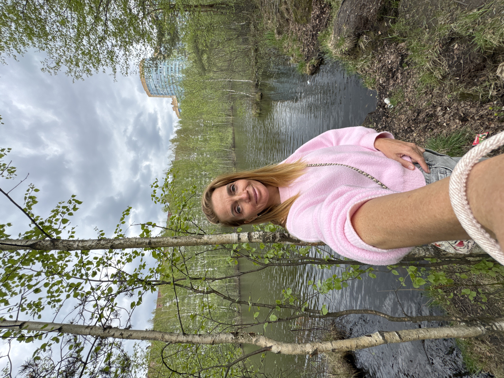 A woman wearing a pink sweater stands in front of a body of water, holding the camera for a selfie. The background includes trees with budding green leaves and a modern building in the distance. The scene is in Saint Petersburg, Russia.