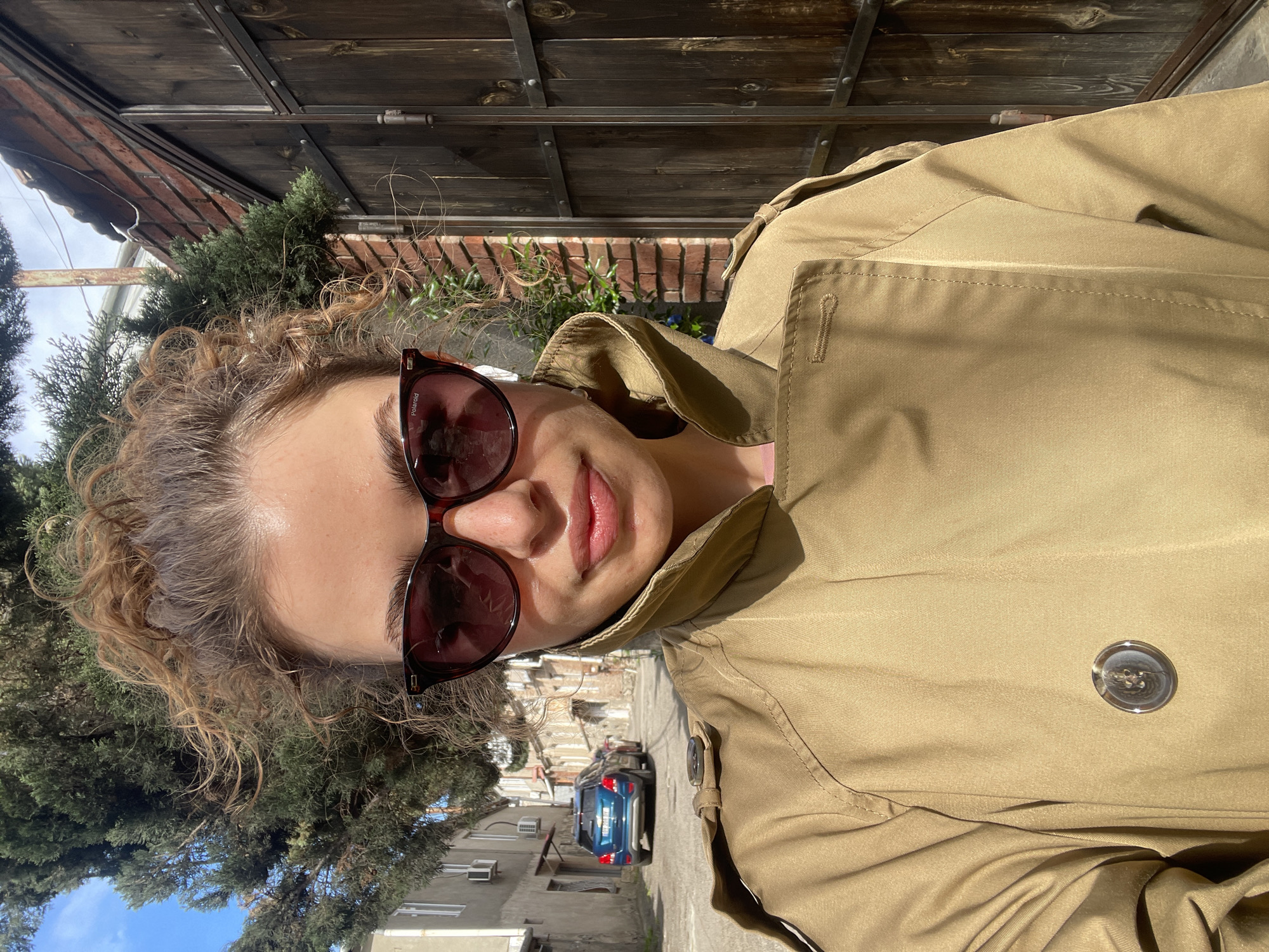 A woman with curly hair and sunglasses is wearing a trench coat. She is standing on a street in Tbilisi, Georgia. Behind her is a wooden gate and a brick wall. In the background, there is a blue car parked on the street. Buildings and trees are visible in the distance. The weather appears sunny.