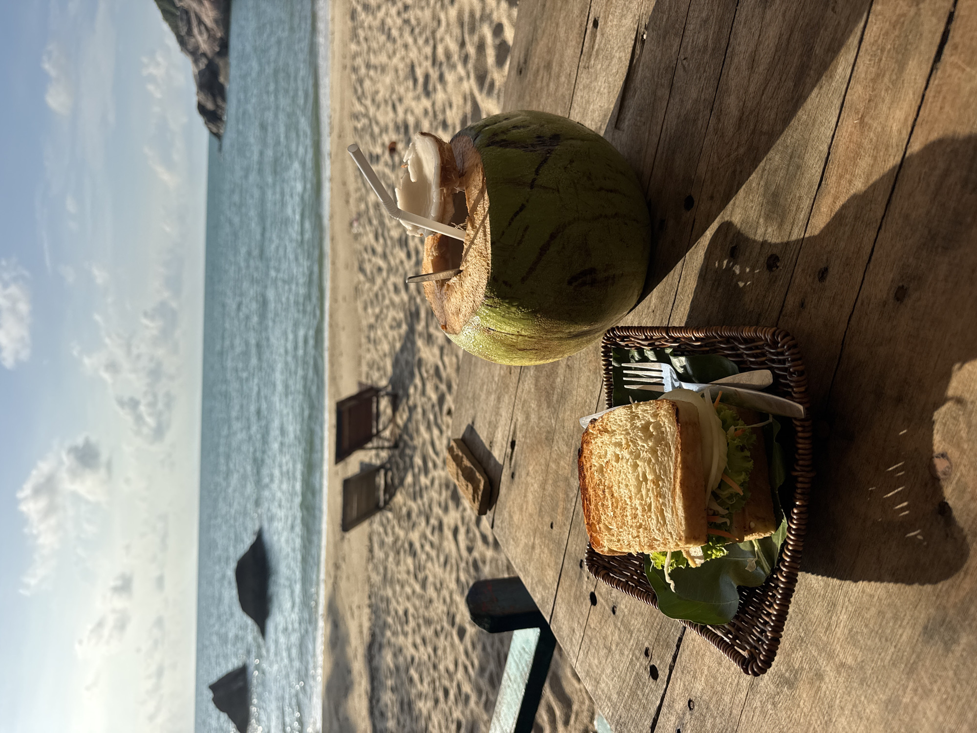 On a wooden table, a cut-open green coconut with straws sits next to a sandwich on a woven plate. The table is located on a sandy beach, with the ocean and sky visible in the background. Two beach chairs are present on the sand. The scene is in Baan Thong Nang, Thailand.