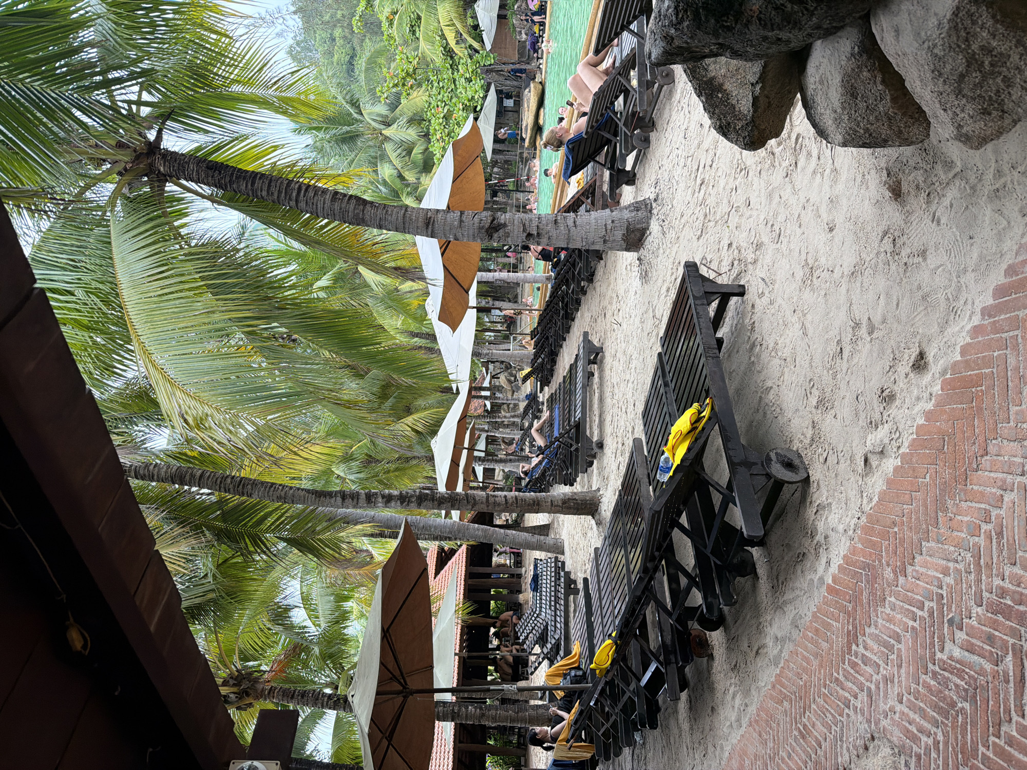 This image depicts a tropical resort setting, likely in Nha Trang, Vietnam, characterized by a sandy beach, numerous palm trees, and a swimming pool. In the foreground, several rows of empty dark wooden lounge chairs are arranged on the sand, some with yellow towels draped over them. A brick pathway runs along the left side of the image. In the mid-ground, people are visible lounging on chairs under large umbrellas and in the vibrant turquoise swimming pool. The pool area is surrounded by lush greenery and more palm trees. In the background, buildings with red tiled roofs suggest the presence of resort facilities. The sky is overcast, indicating a cloudy day. The overall impression is one of relaxation and leisure in a tropical environment.