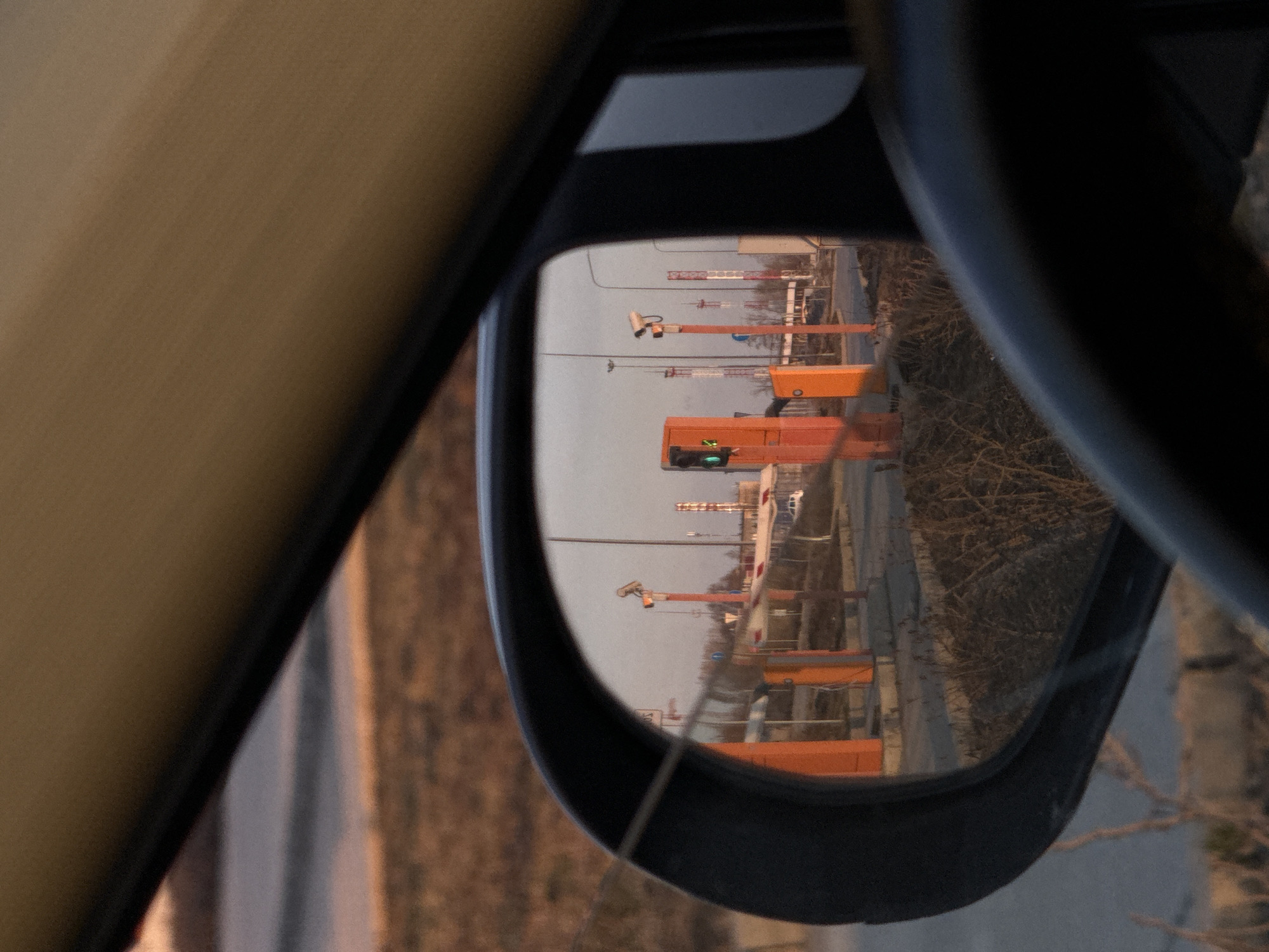 The image shows a car's side mirror reflecting a scene of road construction in Bolshoye Savino, Russia. The reflection captures orange barriers and traffic lights, one of which is green. Construction cranes are visible in the distance. The immediate environment around the construction includes bare trees and infrastructure elements. The photograph is taken from inside a vehicle, evident from the interior paneling and the side mirror's frame.
