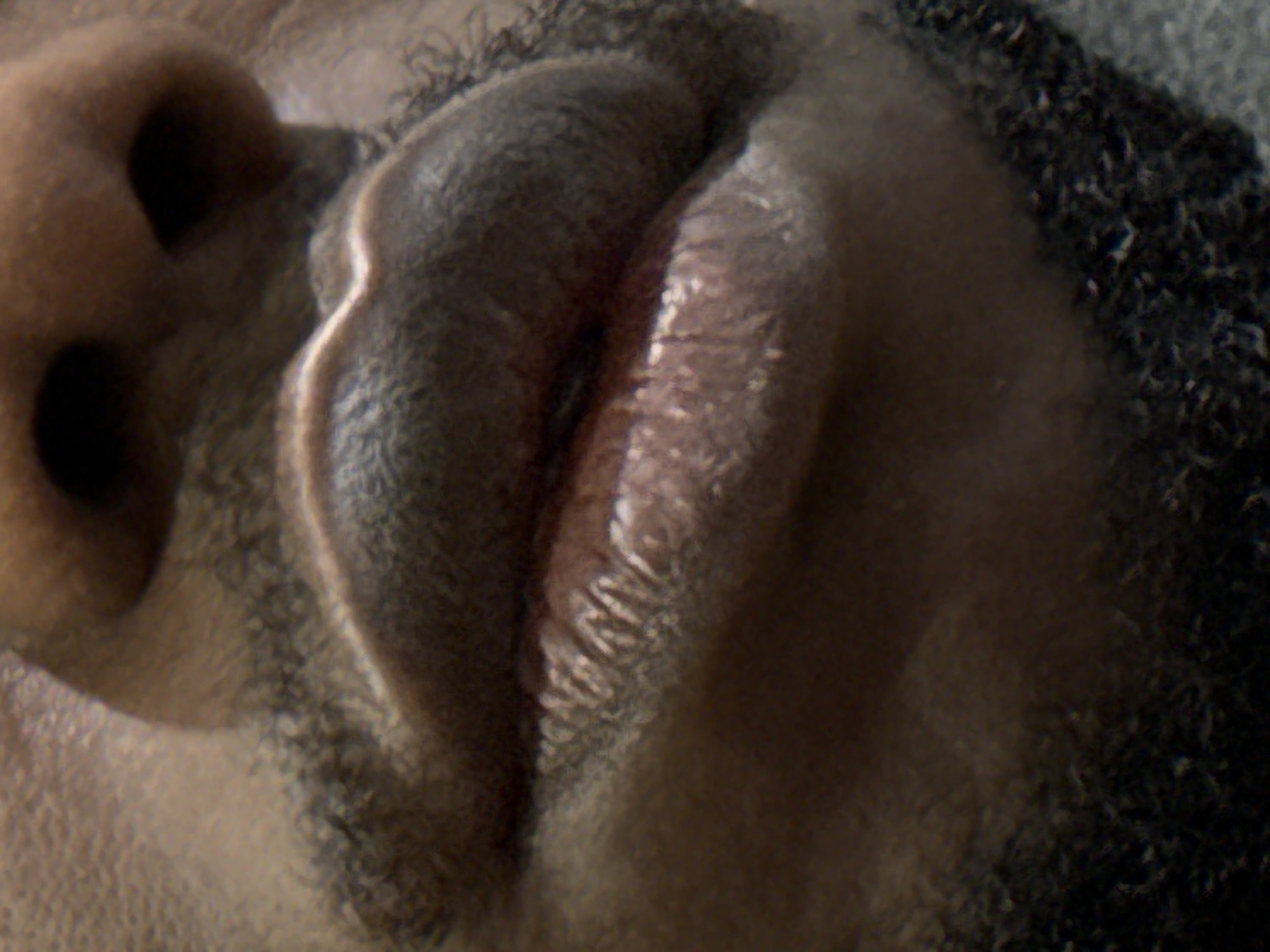 A very close-up, angled portrait shot captures the lower face of a person, likely in Iddo Tudunwada, Nigeria. The image focuses on the nose, mouth, and surrounding facial hair. The subject has dark skin and prominent stubble or short beard growth around the jawline and mouth. The nose is visible on the upper left, with defined nostrils. The lips are full, slightly parted, and appear relaxed, possibly indicating sleep or deep rest. Soft lighting highlights the texture of the lips and skin, creating subtle shadows that give depth to the facial features. The angle of the shot looks upward towards the person's face, emphasizing the contours of the mouth and chin area. No specific activity beyond resting or sleeping is discernible, and no objects or text are visible in the frame. The overall impression is one of intimacy and a focus on the tactile details of the human form.