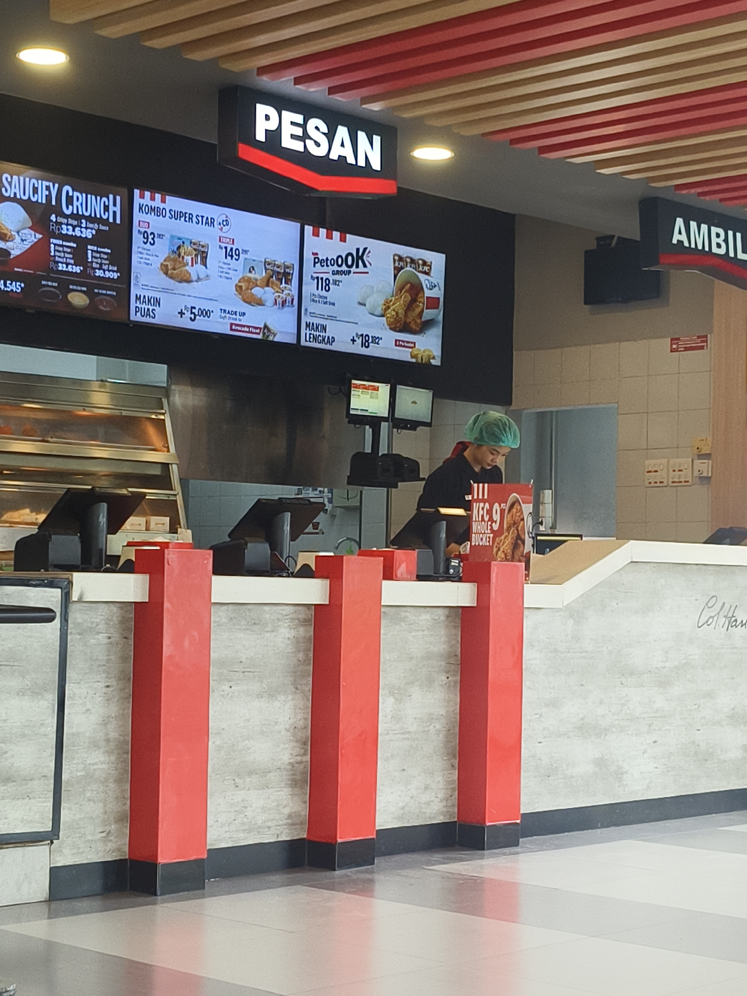 The image shows the interior of a KFC restaurant in Denpasar, Indonesia, with a view of the order counter and menu boards. Above the counter, large digital menu boards display promotional offers and menu items. One board reads "SAUCIFY CRUNCH" with prices, and another displays "KOMBO SUPER STAR" and "PetoOOK". Further to the right, a sign reads "AMBIL". A young woman wearing a green hair net is visible behind the counter, appearing to be an employee. She is standing near a register and a KFC display for a "WHOLE BUCKET". Red and white structural pillars are prominent in the foreground, suggesting an indoor seating area or queueing space. The overall environment is clean and modern, typical of a fast-food chain.