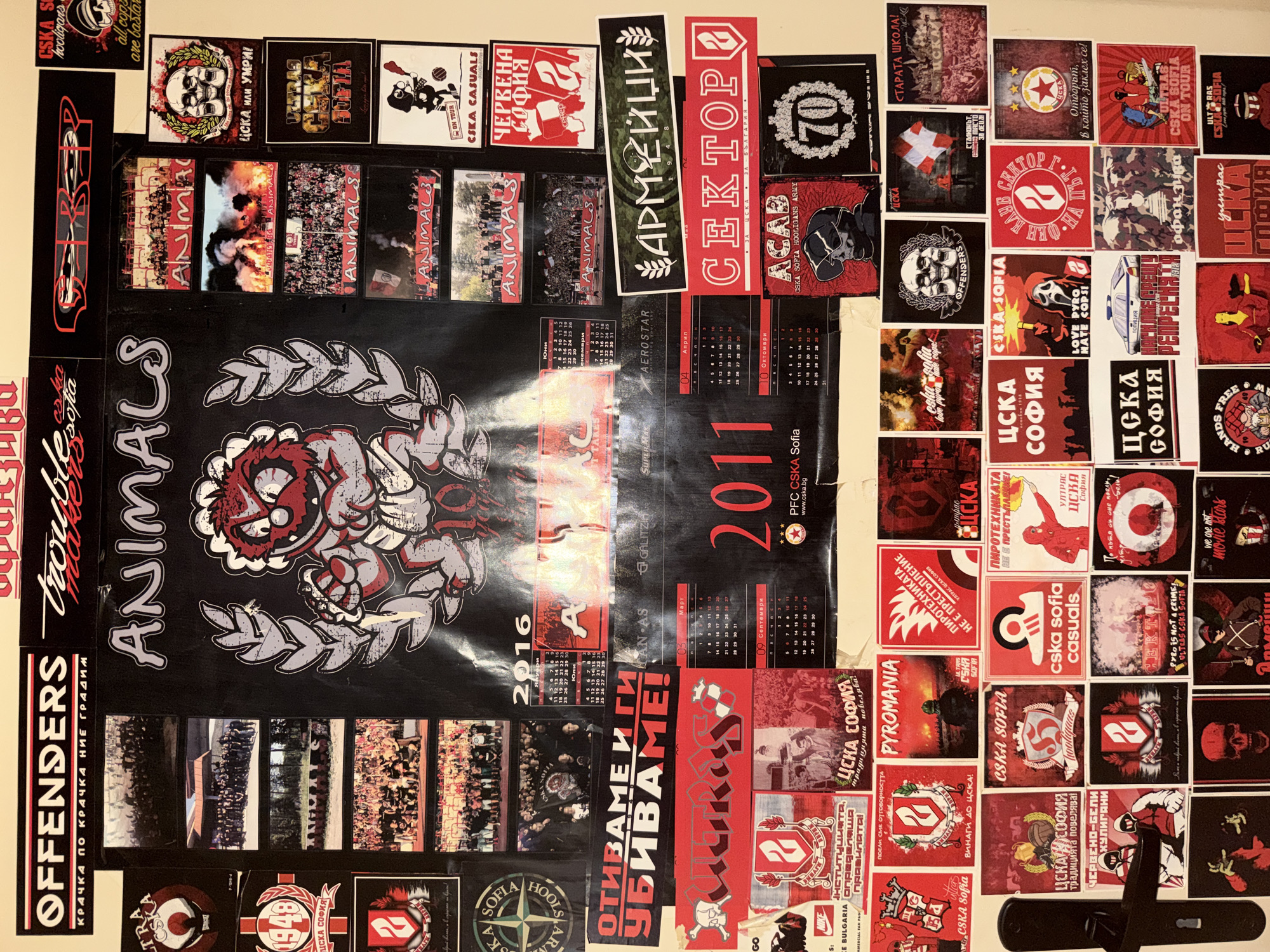 The image displays a wall covered in a dense collage of football fan memorabilia, predominantly stickers and posters related to CSKA Sofia, a Bulgarian football club. The items are arranged in a haphazard manner, creating a chaotic yet visually striking effect. Many of the stickers feature club crests, team slogans in Bulgarian, images of fans, skulls, and pyrotechnics. The colors red and black are heavily featured, consistent with the club's traditional colors. Some of the larger pieces include a "CSKA ANIMALS" poster with a cartoonish character and a calendar for the year 2016. The overall impression is that of a dedicated fan's shrine, possibly located in an apartment or a dedicated fan zone. The door handle of a light-colored door is visible on the left side of the image, suggesting the location is indoors. The lighting appears to be artificial, casting some shadows on the wall.
