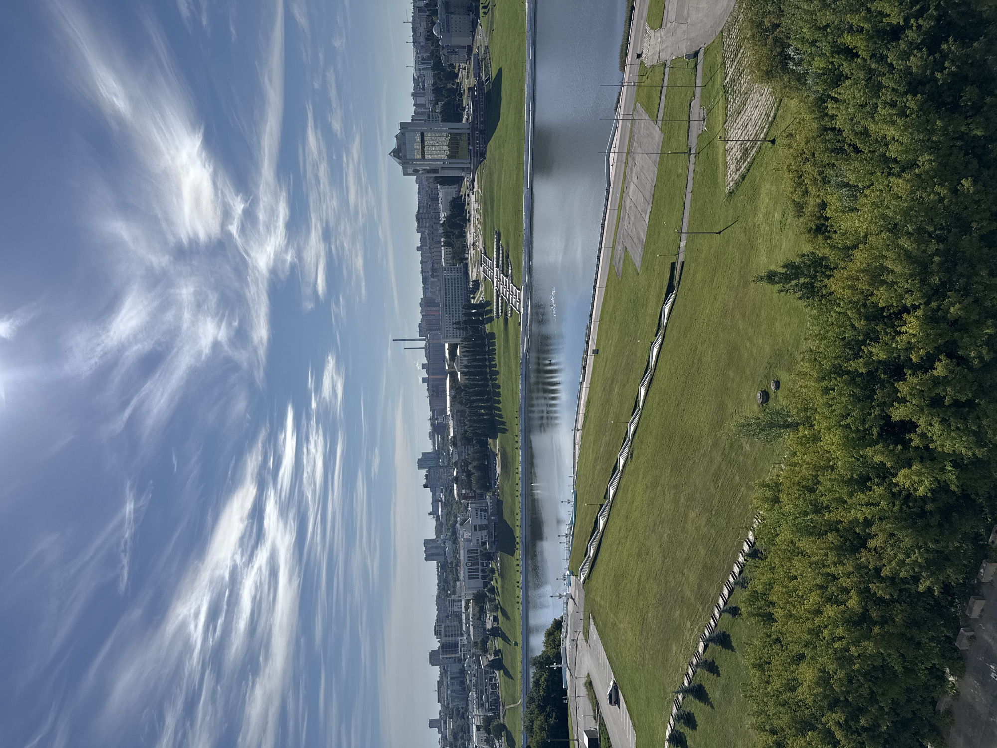This image captures a panoramic view of the Cheboksary Bay in Cheboksary, Russia, on a sunny day with scattered clouds. The foreground is dominated by a lush, green hillside covered in grass and trees. A paved path runs along the edge of the hill, and several light posts are visible.
In the middle ground, a wide river, likely the Cheboksary Bay, reflects the sky and the surrounding cityscape. A white balustrade with ornate lamps lines the riverbank. Further back, a wide expanse of neatly manicured lawn stretches towards the city skyline. This lawn features a cascading water feature or amphitheater with steps, designed to create a striking visual element.
The cityscape in the background showcases a mix of modern and older architecture. Prominent buildings include a distinctive glass skyscraper with a multifaceted design and several other multi-story structures. A large, industrial-looking chimney or tower is visible in the distance. The sky is a vibrant blue, adorned with wispy white clouds that stretch across the frame, illuminated by the bright sun. The overall scene conveys a sense of urban development integrated with natural and man-made recreational spaces. No people are discernible in the image.