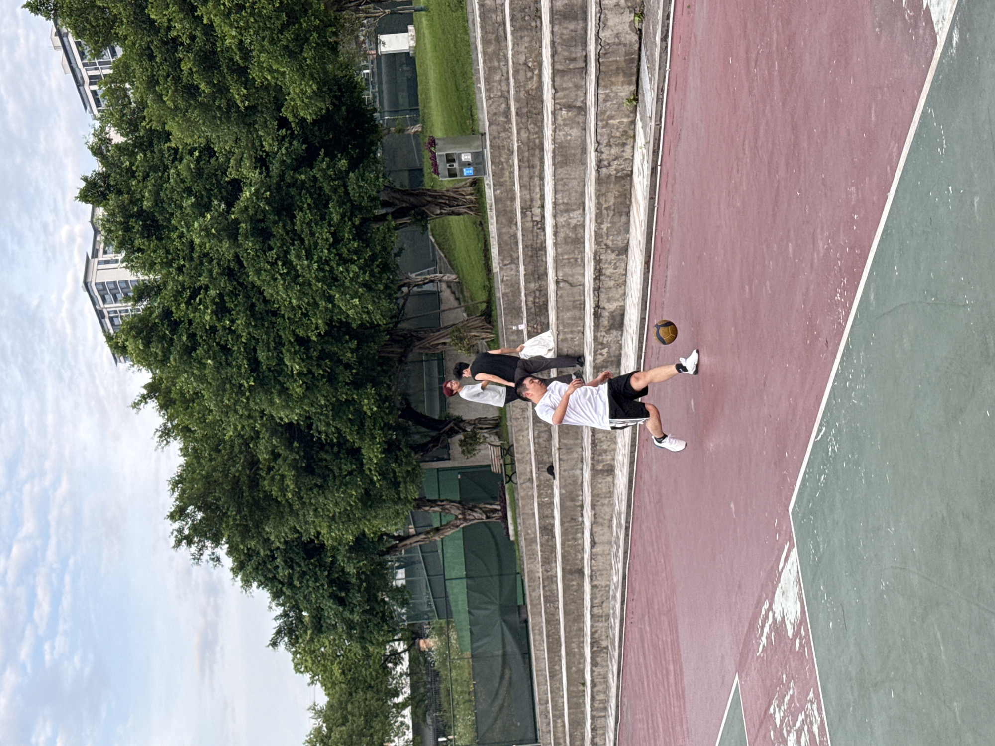A full shot depicts a man in sportswear kicking a soccer ball on a partially green, partially red concrete court. The court is bordered by concrete steps on one side and mature trees and green mesh fencing on the other. Two other people are sitting on the steps. The left of the image features a partially visible apartment building. The sky is overcast. There is no explicit indication of this image being located in Jiangjin, China.