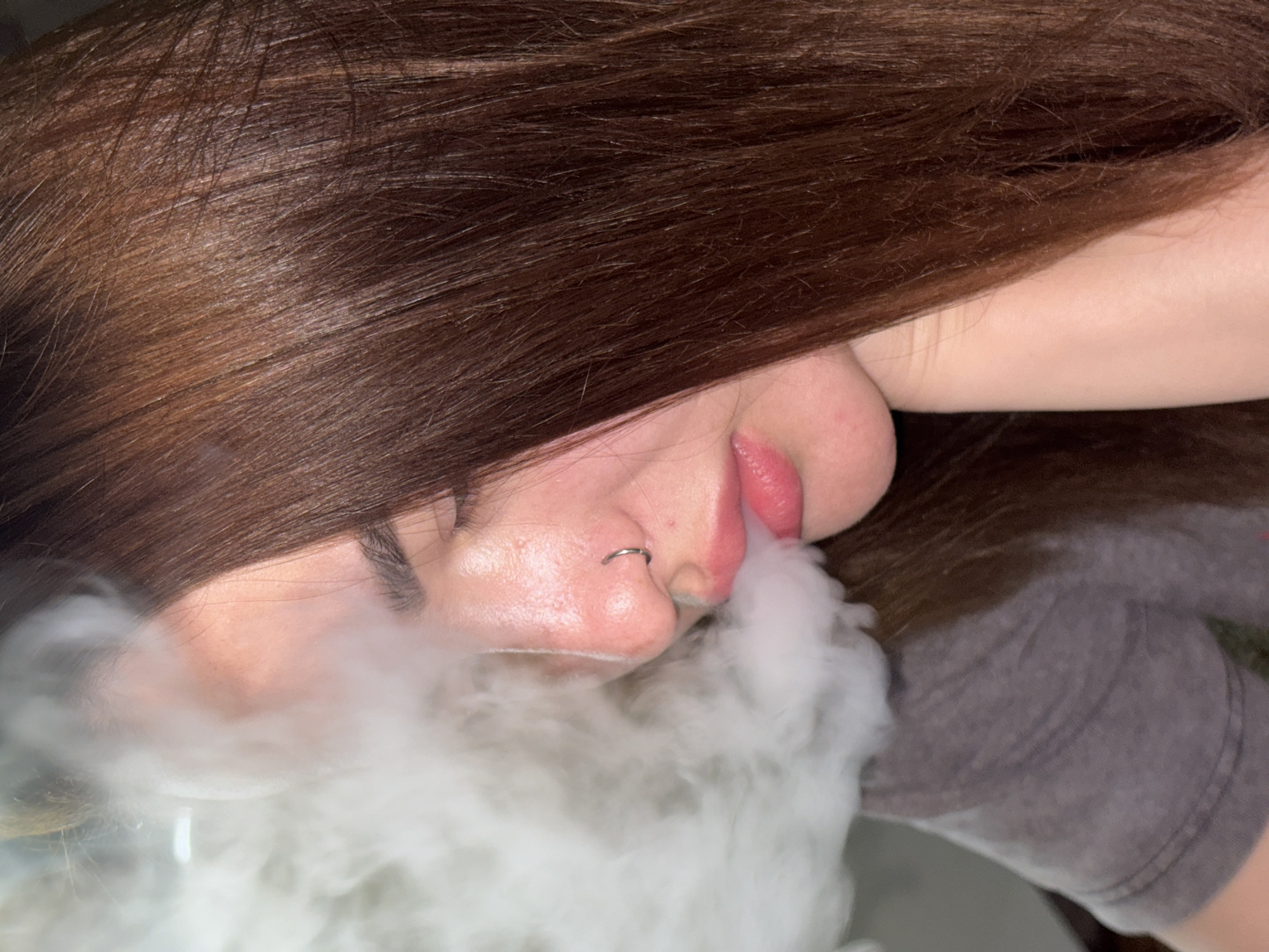 A close-up, angled shot depicts a person exhaling a large cloud of vapor. The individual has brown hair that partially obscures their face. A silver ring is visible in their left nostril. The person is wearing a gray t-shirt. The location is not discernible from the provided image.