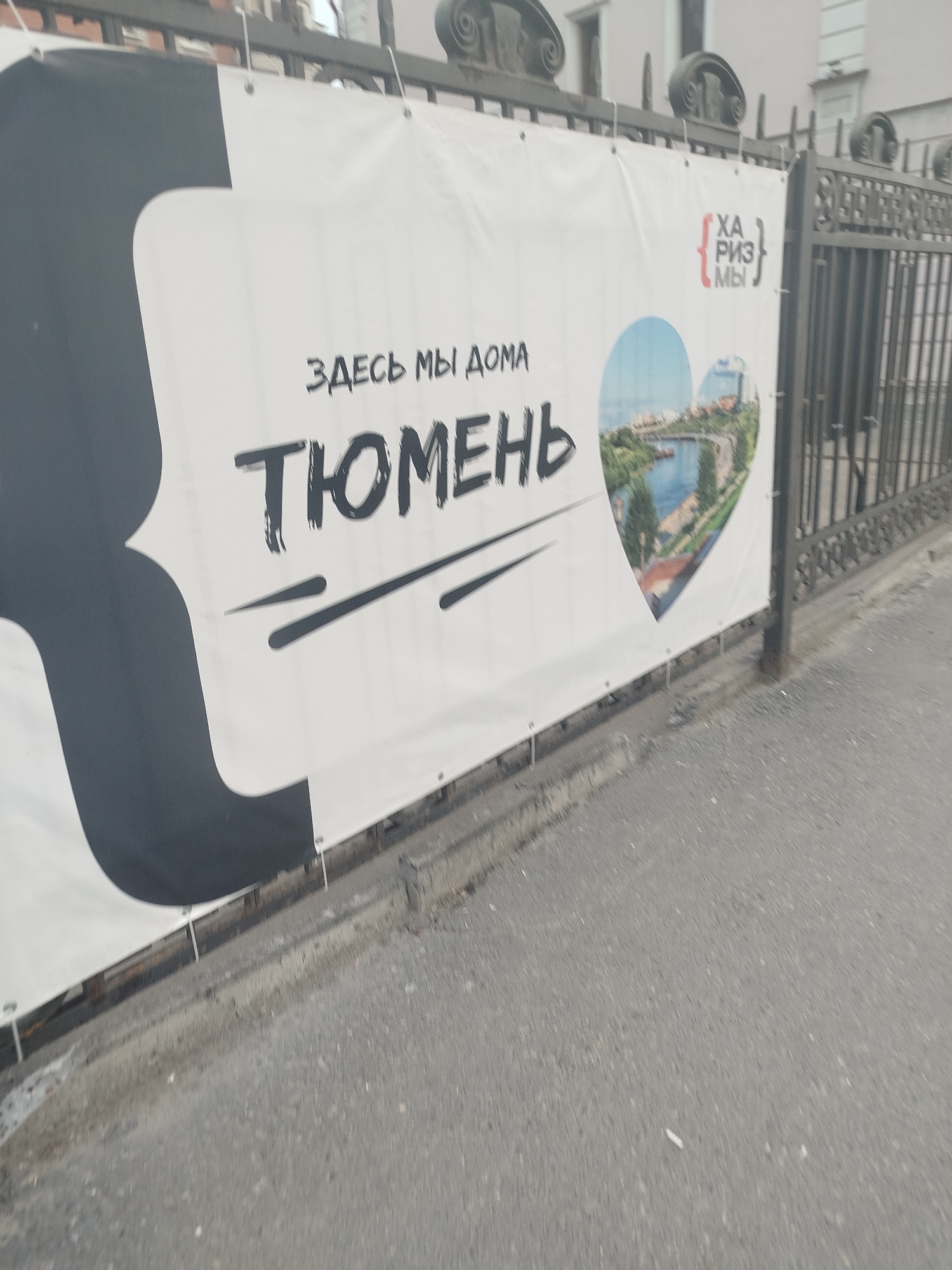 The image shows a large white banner with black and red text and a heart-shaped graphic featuring a cityscape. The text on the banner reads "ЗДЕСЬ МЫ ДОМА" (Here we are home) and "ТЮМЕНЬ" (Tyumen). To the right of the text is a red graphic with curly brackets and the word "ХАРИЗМЫ" (Charisma). The heart-shaped graphic depicts a river and buildings. The banner is attached to a black metal fence. The scene is outdoors, with an asphalt ground in the foreground. The banner appears to be hung across a street or walkway, possibly as a decorative element or advertisement related to the city of Tyumen, Russia.