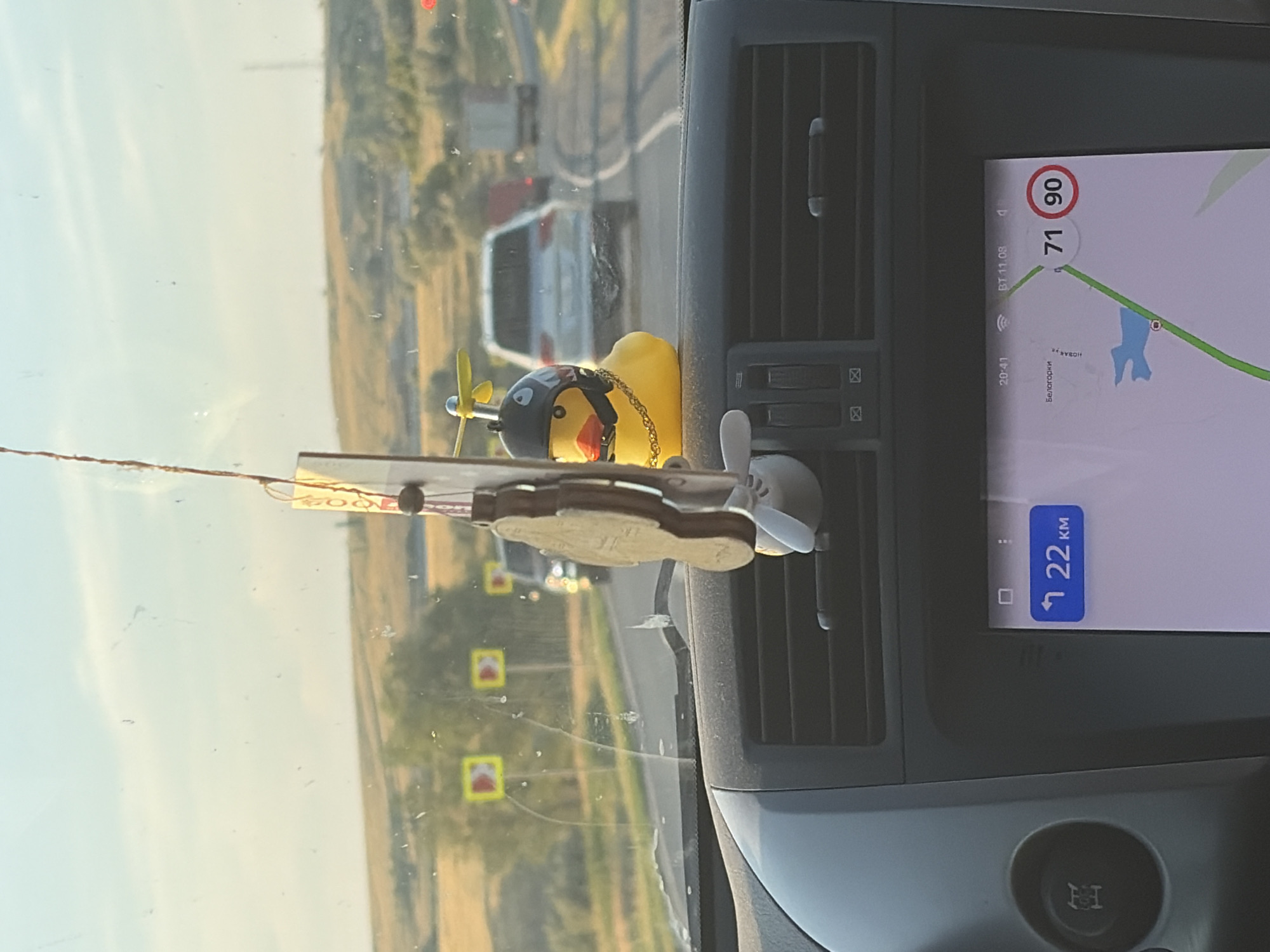 The image, taken from inside a vehicle, displays a dashboard with a navigation screen showing a map of Belogorki, Russia. The screen indicates a speed limit of 71 out of 90 and a distance of 22 km to the destination. A yellow rubber duck with a pilot helmet and propeller is attached to the rearview mirror as an air freshener. Outside the vehicle, a highway is visible with several cars, trucks, and road signs. The sky is clear and the sun is setting, casting a warm light on the landscape.