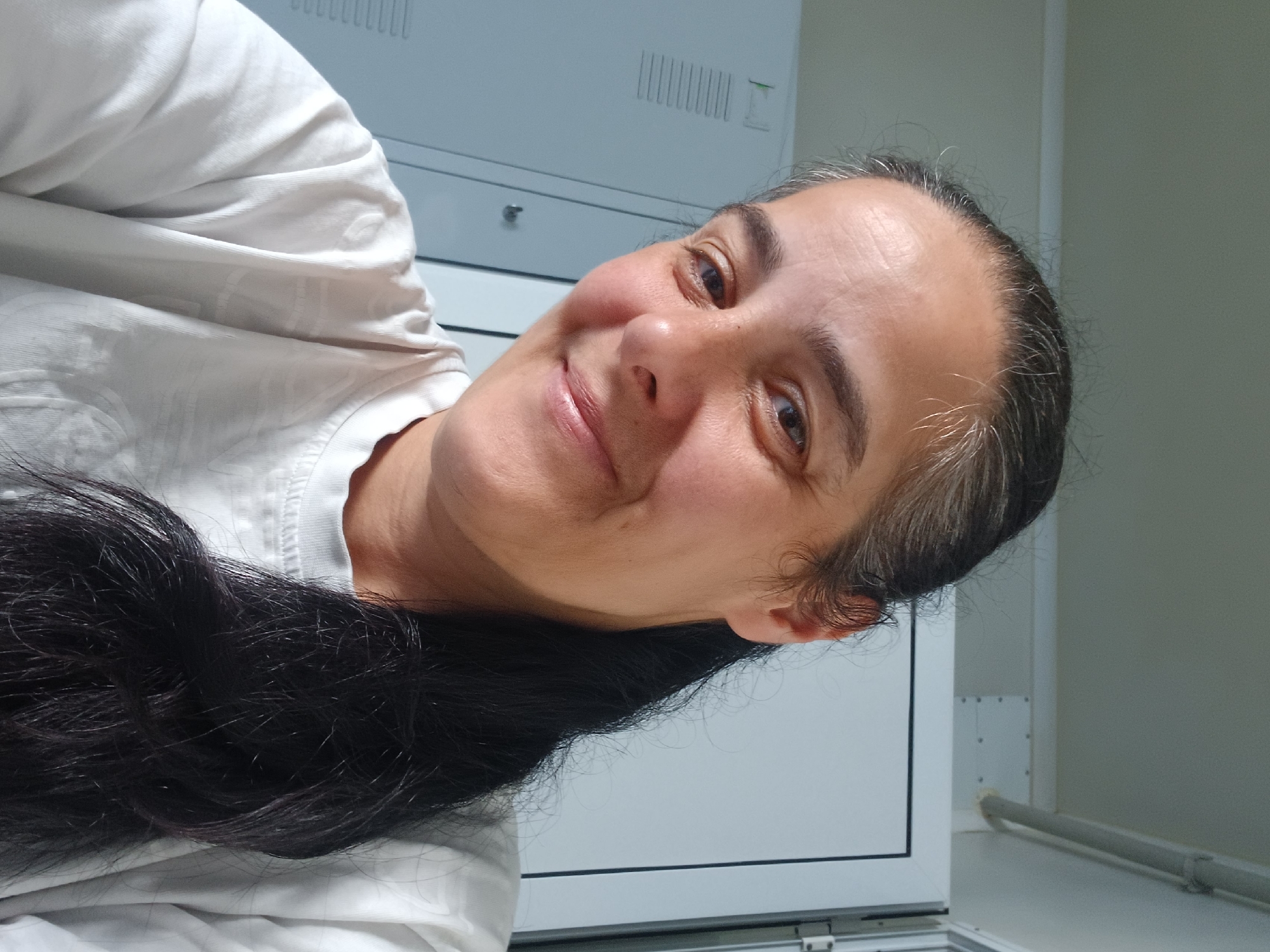 A medium shot taken from a slightly high angle shows a woman in her late 30s to early 40s with medium-toned skin and dark hair pulled back into a ponytail that falls over her right shoulder. She has strands of gray hair mixed with the dark hair on her head, and a few stray hairs frame her face. Her eyebrows are dark and well-defined, and her eyes are open, looking directly at the camera. She has a slight smile, with her lips closed. She is wearing a plain white t-shirt with a crew neck. The background is a white, sterile-looking room with what appear to be lockers or cabinets on the left and a white door or panel on the right. The environment suggests an indoor setting, possibly a locker room or changing area. The image is well-lit, with natural light casting soft shadows. The overall impression is that of a casual selfie taken indoors. There is no indication of the city of Rakevo, Bulgaria within the image itself.