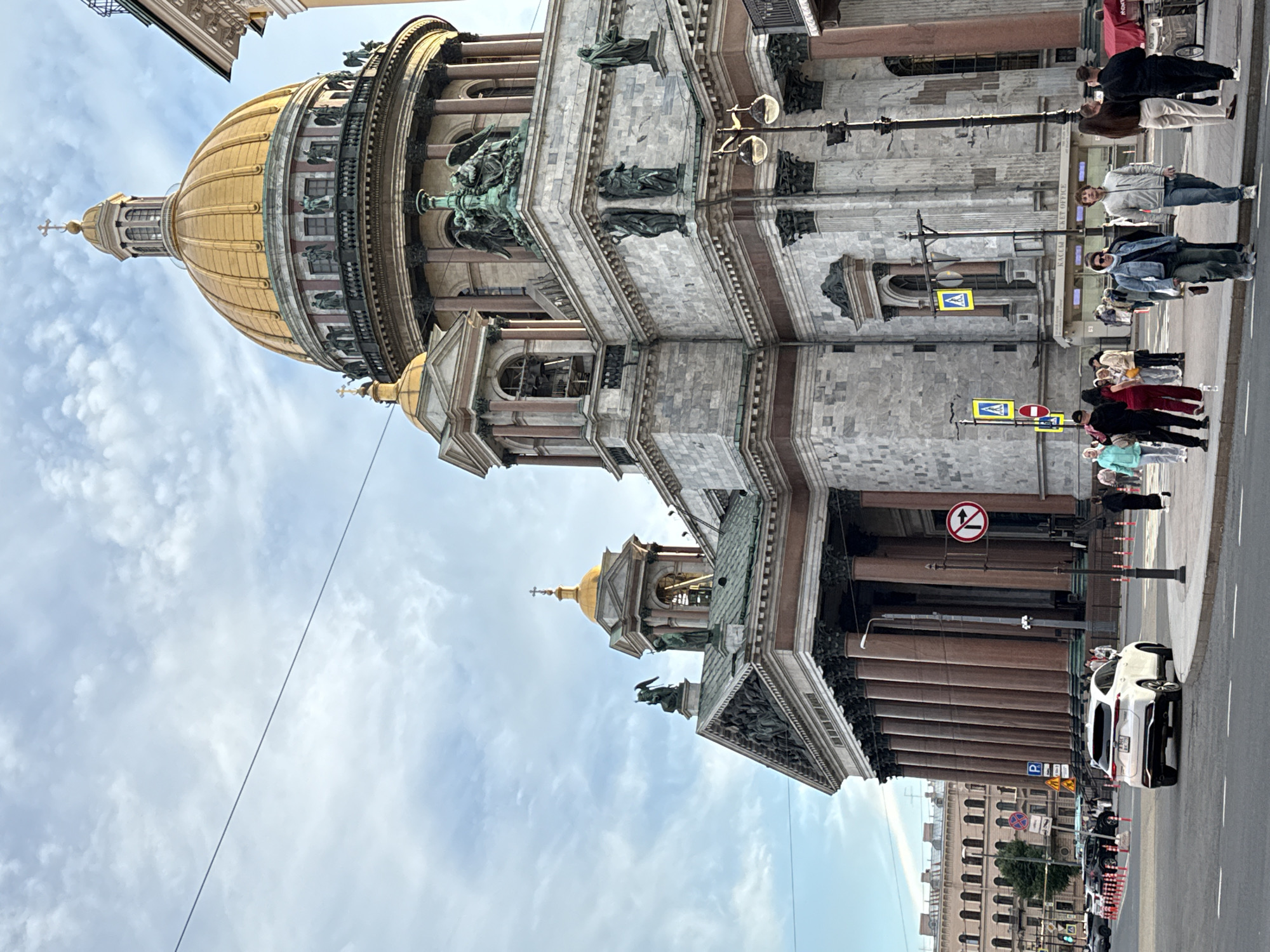 The image depicts the facade of a grand, ornate building, likely a cathedral, with a prominent golden dome topped with a cross. Sculptures adorn the exterior, including figures in niches and a winged statue. The building is constructed of stone and features large columns and detailed architectural elements.
In the foreground, people are walking on a sidewalk and crossing a street. They are dressed in casual clothing, suggesting a daytime outing. Some individuals appear to be tourists, possibly taking in the sights. A white car is parked on the side of the road.
Street signs are visible, including one with directional arrows and a red circle with a diagonal line, indicating a prohibition. Another sign has a blue square with a white 'P', signifying parking. The sky is partly cloudy. The architecture and the presence of people suggest an urban setting, and the information provided indicates the location is Saint Petersburg, Russia. The overall atmosphere appears to be calm and observational, with no overt signs of distress or excitement among the people. The lighting suggests it is daytime, likely late morning or afternoon.