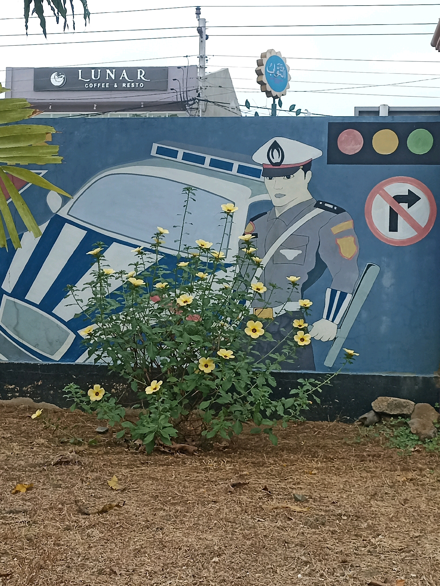 In Panjer, Indonesia, a mural depicts a police officer in uniform with a baton, standing next to a stylized police car. Above the mural, a sign reads "LUNAR COFFEE & RESTO". A traffic signal with red, yellow, and green lights is painted on the wall, along with a "no right turn" sign. In the foreground, a flowering bush with yellow blossoms adds a natural element to the urban scene. The ground is covered in dry grass and fallen leaves.