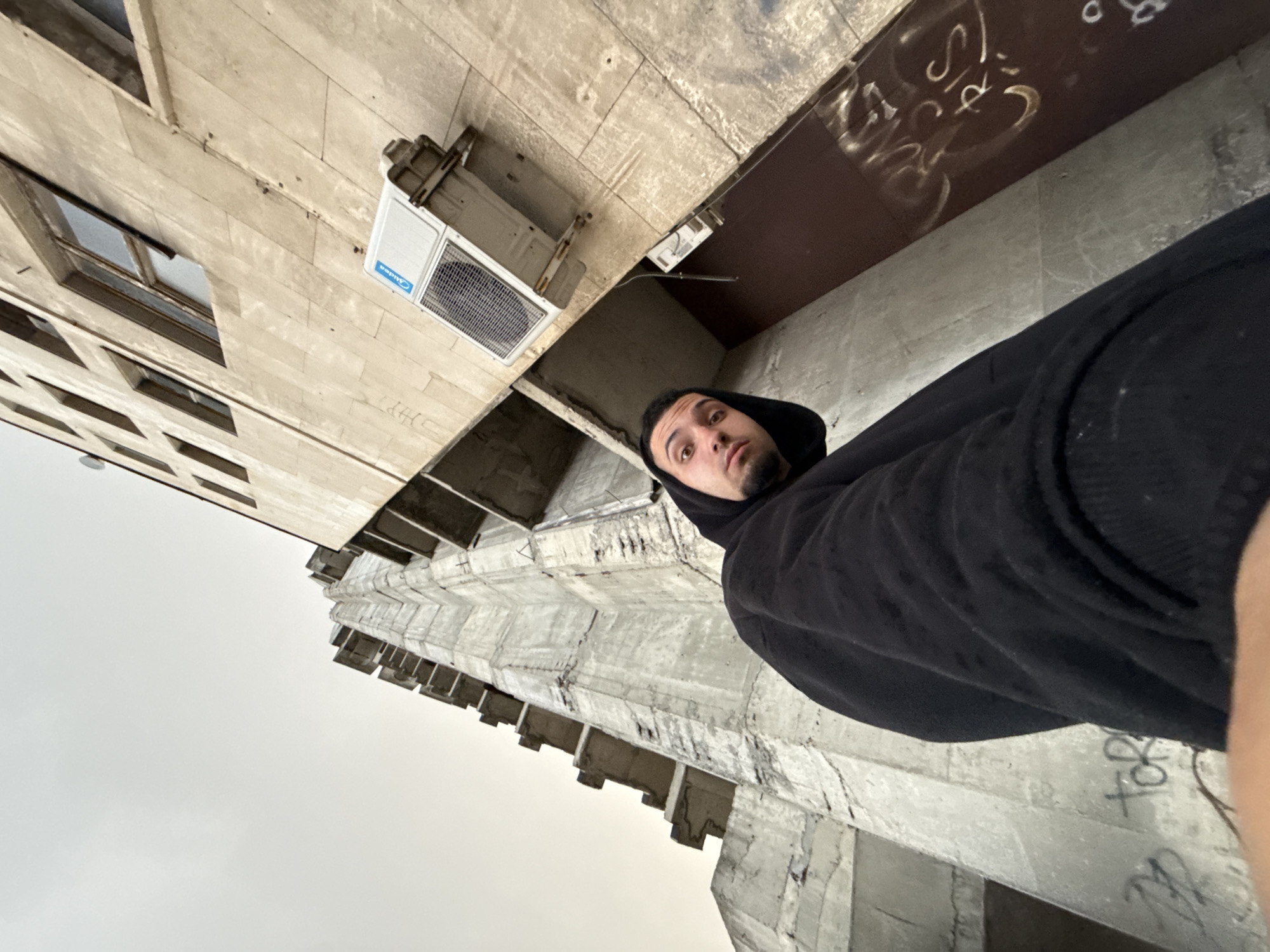 A low-angle shot shows a man in a black hoodie looking into the camera. He appears to be standing near a large, weathered concrete structure. In the background, a tall building is visible, with what looks like an air conditioning unit attached to its side. Graffiti is present on the concrete surfaces. Based on the architectural style and the presence of similar concrete structures, this image likely depicts a scene near the Founders of the Bulgarian State Monument in Shumen, Bulgaria.