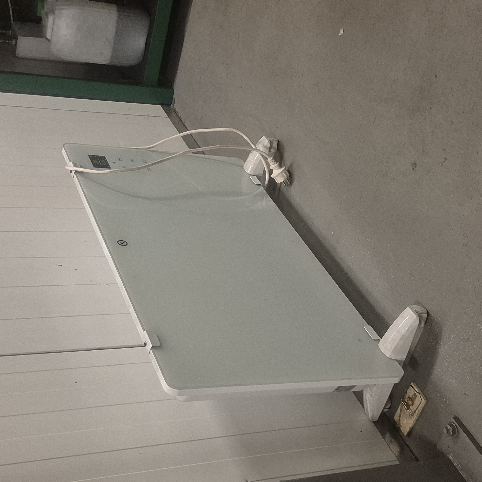 In Nowa Wieś Książęca, Poland, a white electric panel heater stands on a grey concrete floor, positioned against a white, vertically grooved wall. The heater has two white feet and a power cord plugged into an outlet. To the left of the heater, near the wall, a wooden mousetrap is visible on the floor. In the background to the right, a green metal shelf unit holds a large translucent plastic container filled with a grey liquid. The lighting is dim, suggesting an indoor setting, possibly a storage area or utility room.