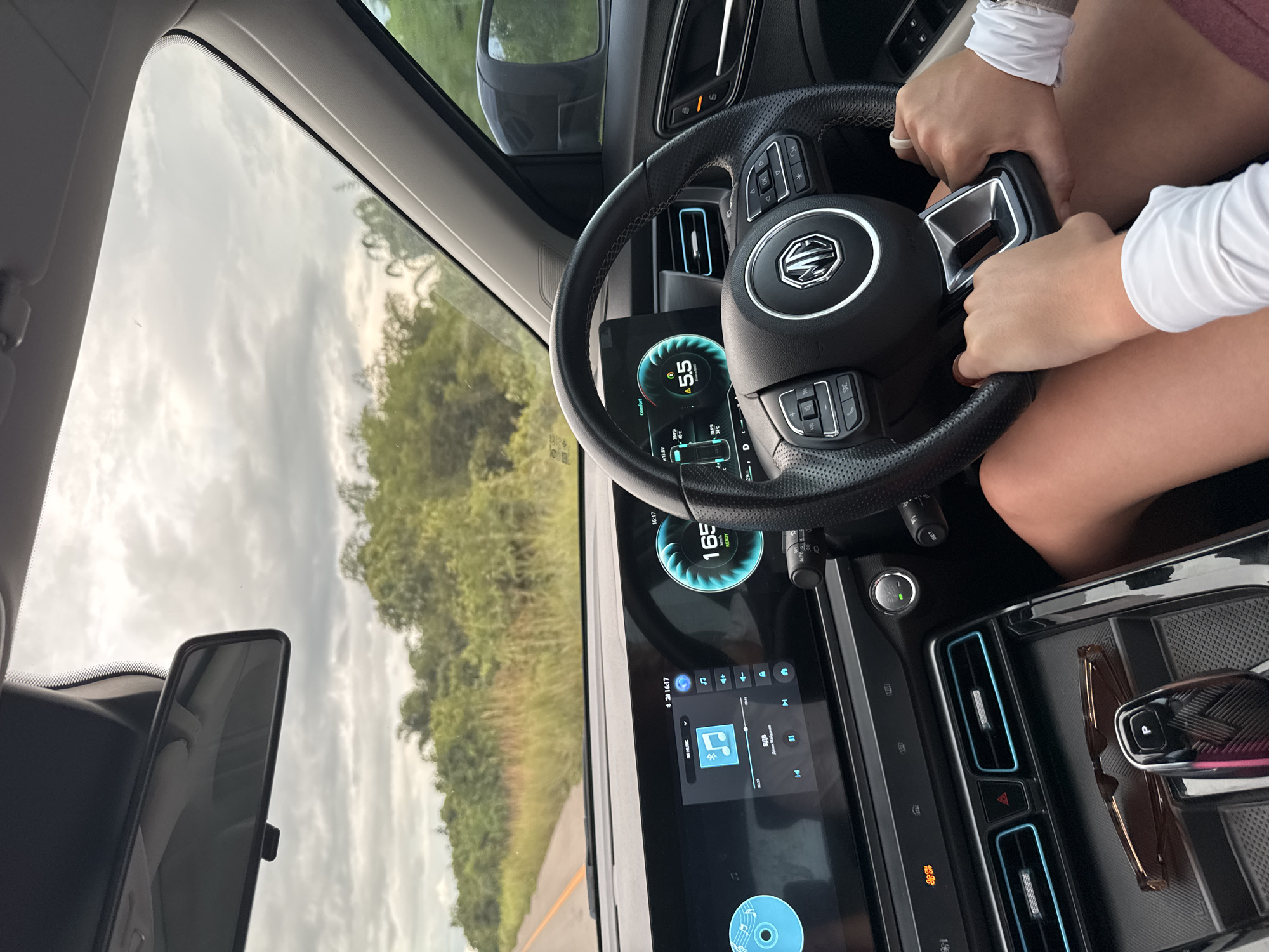 In Plai Phraya, Thailand, a passenger's hands are visible resting on the steering wheel of a car. The driver's hands are also on the wheel, and their legs are visible. The car's dashboard displays include a speed of 165 km/h and a time of 16:17. Outside the car, a road stretches ahead, bordered by lush green vegetation under a cloudy sky. Sunglasses and a car key are visible in the center console.