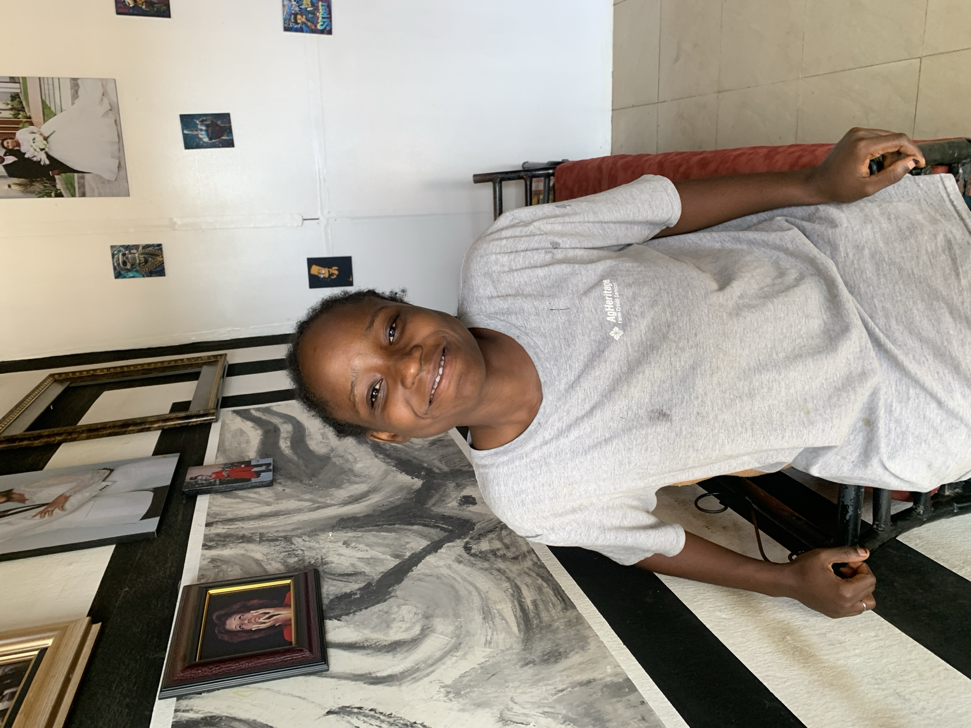 A woman is lying on a padded bench indoors, likely in a residential setting. She is smiling and looking directly at the camera. She is wearing a grey t-shirt with the logo "AgHeritage Farm Credit Services" visible. The floor and wall are decorated with a black and white stripe pattern and an abstract black and white painting. Several framed photos and images are mounted on the wall behind the woman. The location is potentially in Jos, Nigeria, given the request's prompt.