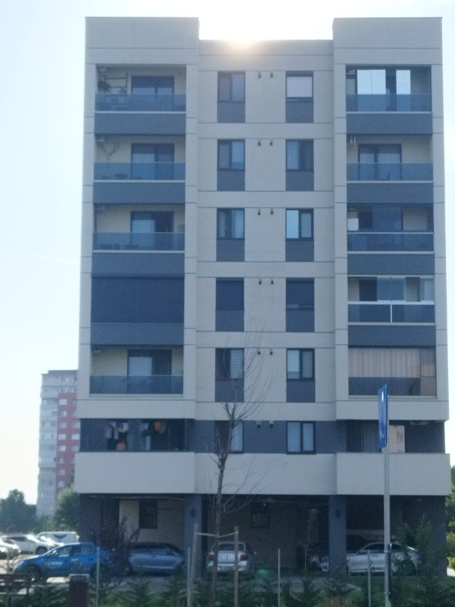 A tall, modern apartment building with a beige and dark grey facade dominates the frame, set against a bright sky with visible sun glare. The building features multiple floors with balconies and windows. The balconies have blue glass railings. On one of the balconies, clothes are hung out to dry. Below the main structure, the building has an open ground floor used as parking, with several cars parked there. In the foreground, more cars are visible, including a blue sedan and a silver hatchback, parked on a paved area. Trees and greenery surround the parking area. To the left, another, older apartment building with a red and white facade is partially visible. A blue street sign on a pole stands near the right edge of the apartment building. The scene appears to be a residential area in Bucharest, Romania, likely during daylight hours.