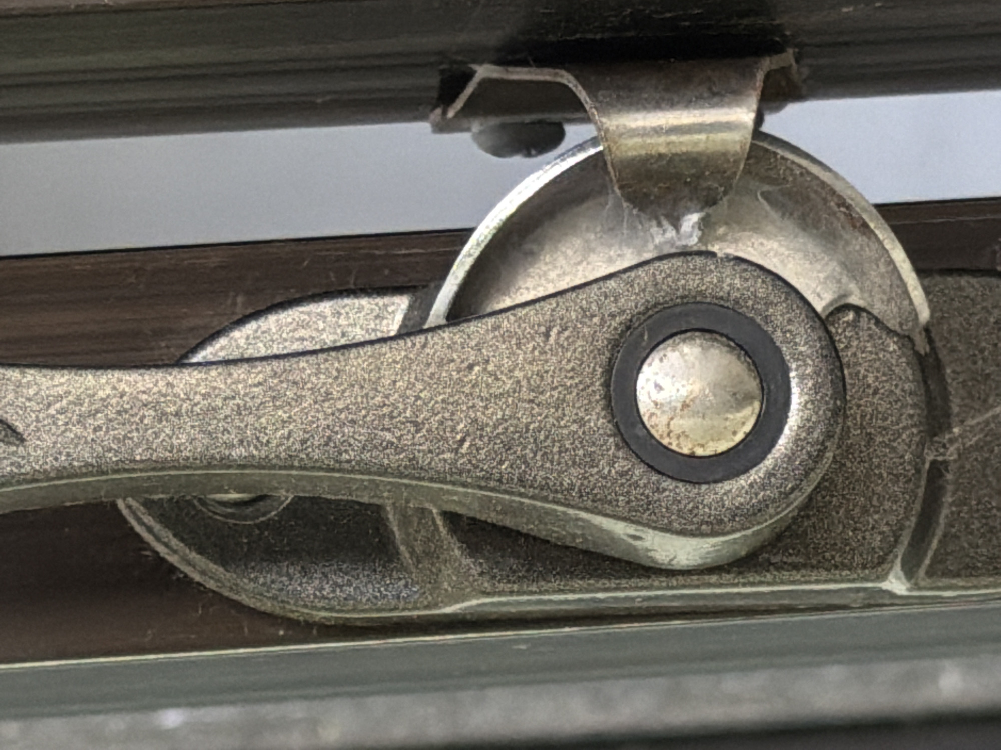 The image presents a tight close-up of a window operating mechanism, likely part of a casement or awning window. The central subject is a textured, dark silver-grey metallic latch or crank, showing signs of use and dust accumulation. This mechanism consists of an elongated handle connected to a circular pivot point, which in turn engages with a larger, gear-like circular component.
Above the main rotating part, a smaller, shiny metal clip or bracket is visible, secured by a screw to the upper section of a dark brown or black window frame. Fine dust particles and strands resembling cobwebs are clearly visible on the metallic surfaces, particularly on the larger rotating wheel and within the crevices of the mechanism, suggesting it hasn't been recently cleaned.
The background reveals a lighter, muted grey area, which could be the glass pane of the window or a surface beyond it. The surrounding window frame itself is a dark, possibly wooden or metallic, material. No people are present, and no activity is depicted; the mechanism is stationary, implying its function to open or close a window. The lighting is ambient and even, offering no specific cues for the time of day. There is no visible text on any of the components. The scene offers a detailed view of everyday window hardware, consistent with a setting in North Kuta, Indonesia.