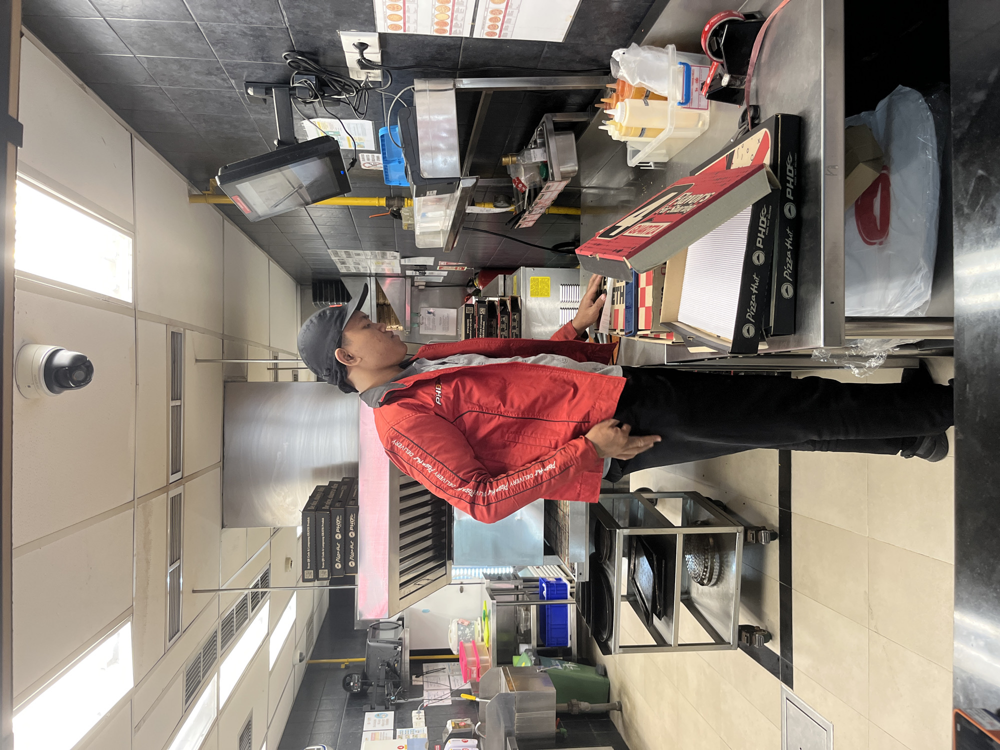A male employee wearing a red Pizza Hut jacket and a black baseball cap is standing in a commercial kitchen, preparing pizza boxes. He is looking down at the boxes and appears to be in the process of assembling or organizing them. The kitchen is equipped with stainless steel countertops, a commercial-grade stove hood, shelves filled with supplies, and various food preparation equipment. The walls are tiled with black and white tiles. A security camera is visible on the ceiling. The overall environment suggests a busy food service establishment, likely a Pizza Hut restaurant, possibly in Kuta Selatan, Indonesia, given the context.