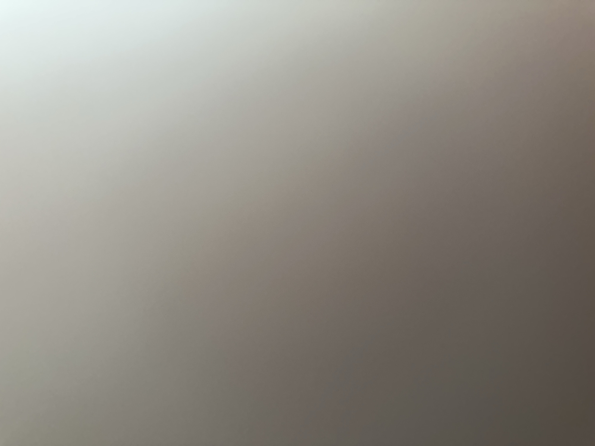 This is a photograph of a plain, neutral background, likely an indoor surface. The lighting is soft and diffused, creating a gradient from a brighter, lighter area in the upper left to a darker, more muted tone towards the lower right. There are no discernible subjects, objects, or any activity taking place. The image lacks specific details about the time of day, weather, or location. No text is visible. The overall impression is one of abstract texture and light.