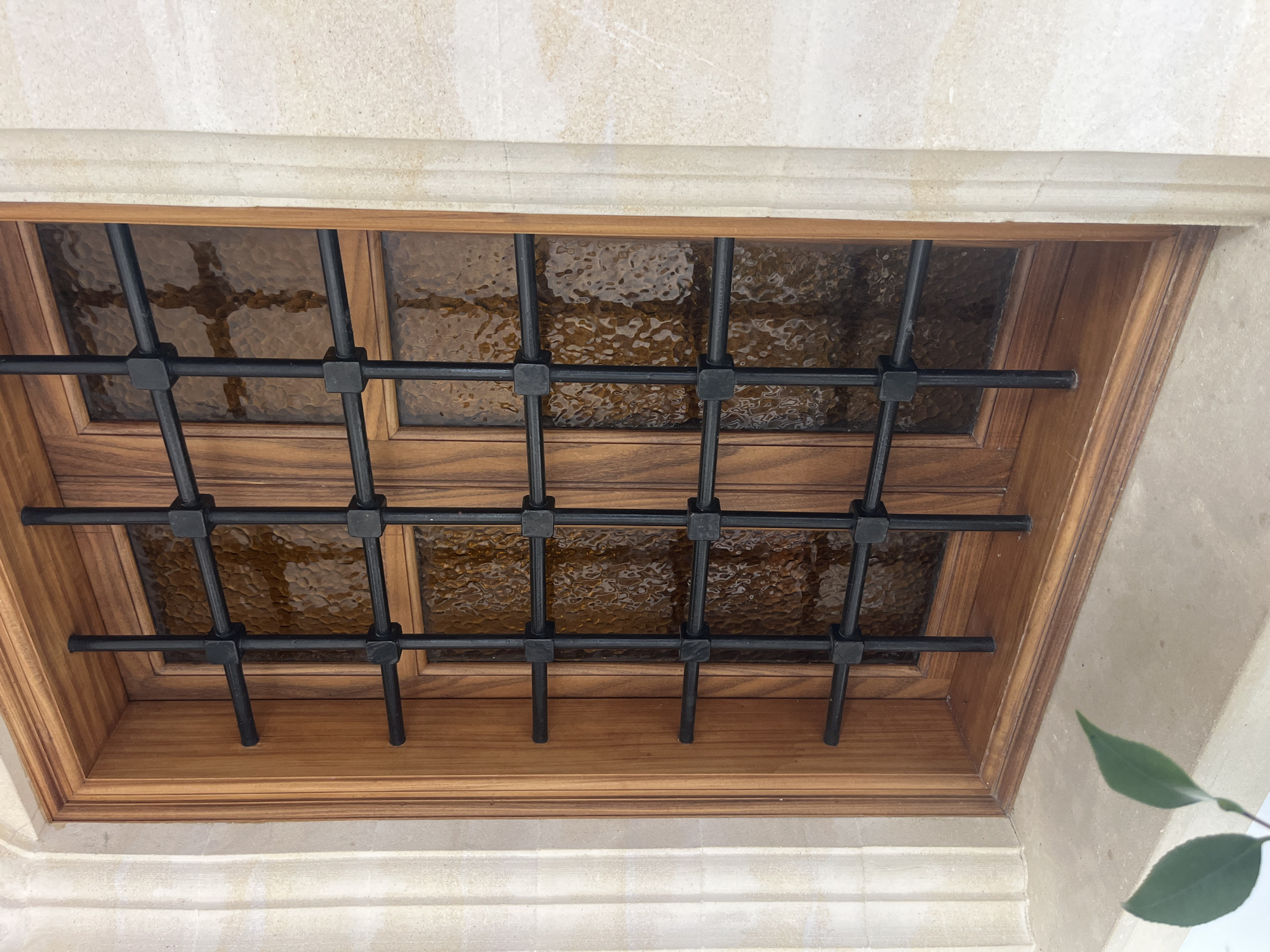 This is a medium shot of a window set into a light beige stone wall. The window frame is made of polished dark wood, and it contains two panes of textured brown glass. A black metal grate with a grid pattern is affixed to the exterior of the window. The grate has vertical bars and horizontal bars, with square connectors at each intersection. The lower sill of the window is also made of the same light beige stone as the wall. Two green leaves from a plant are visible in the lower left corner of the image, resting on the stone sill. The scene appears to be an exterior detail of a building in Foini, Cyprus.
