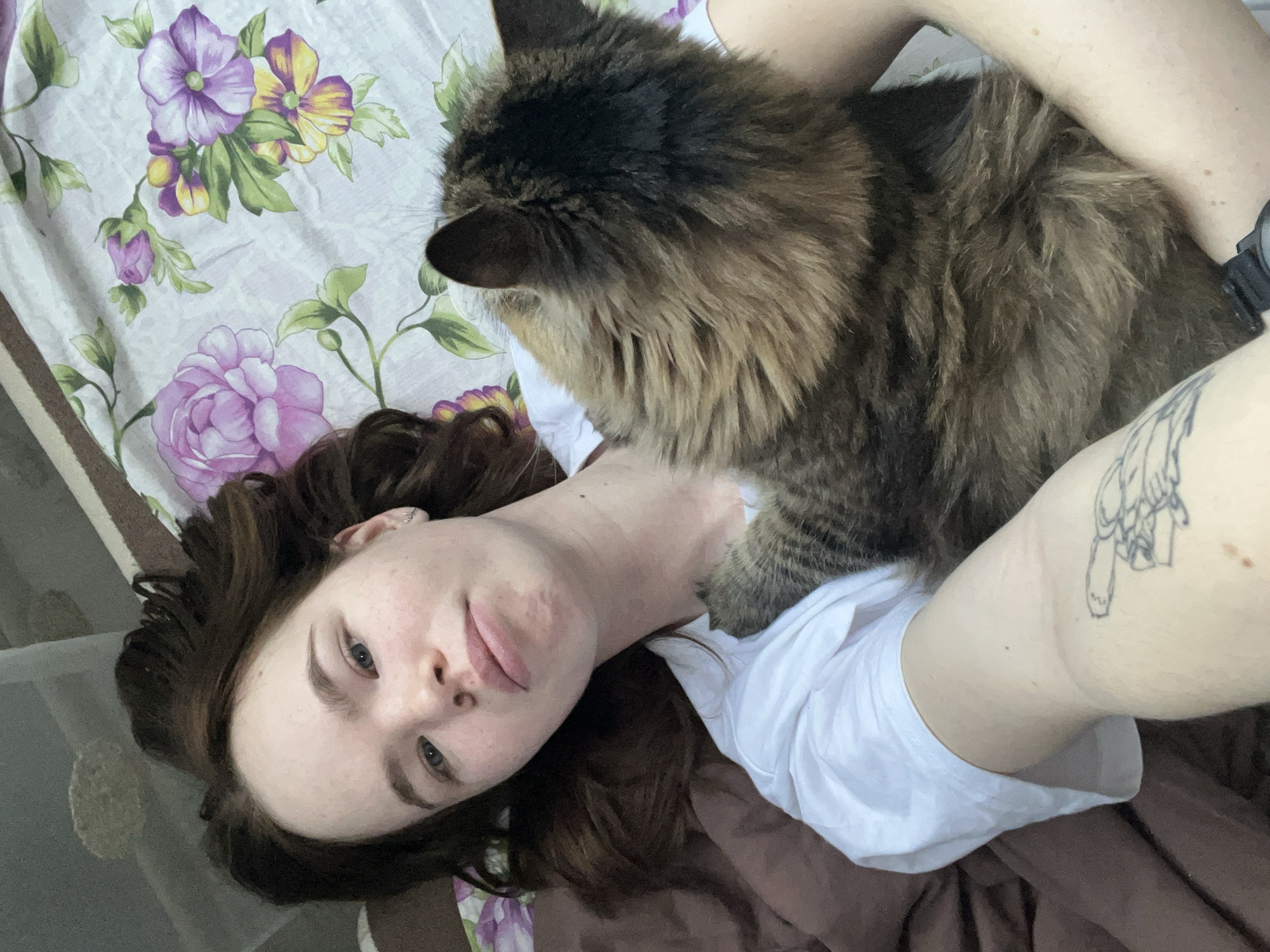 A woman with brown curly hair, light skin, and neutral expression lies on her back in a bed. A large, fluffy brown and black cat rests on her chest. The bed has a floral patterned sheet with purple and yellow flowers. A brown blanket is visible at the bottom of the frame. The woman wears a white t-shirt, a watch on her left wrist, and has a tattoo on her left arm. The location is unknown, and there is no discernible information to suggest the location is Chaikovsky, Russia.