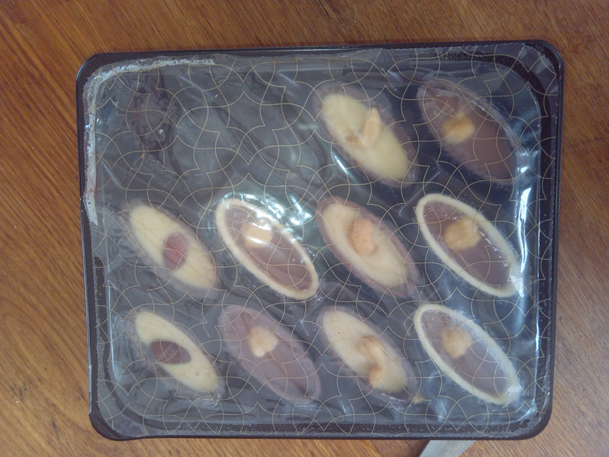 A rectangular, clear plastic package containing twelve oval-shaped chocolates is presented on a wooden surface. The chocolates are arranged in three rows of four. Each chocolate features a lighter-colored topping, possibly marzipan or white chocolate, with a darker chocolate base. Some chocolates have what appear to be nuts or dried fruit embedded in the lighter topping. The packaging is dark with a subtle gold geometric pattern. The wooden surface is a medium brown with visible grain. There is no indication of the location within Terebovlia, Ukraine.