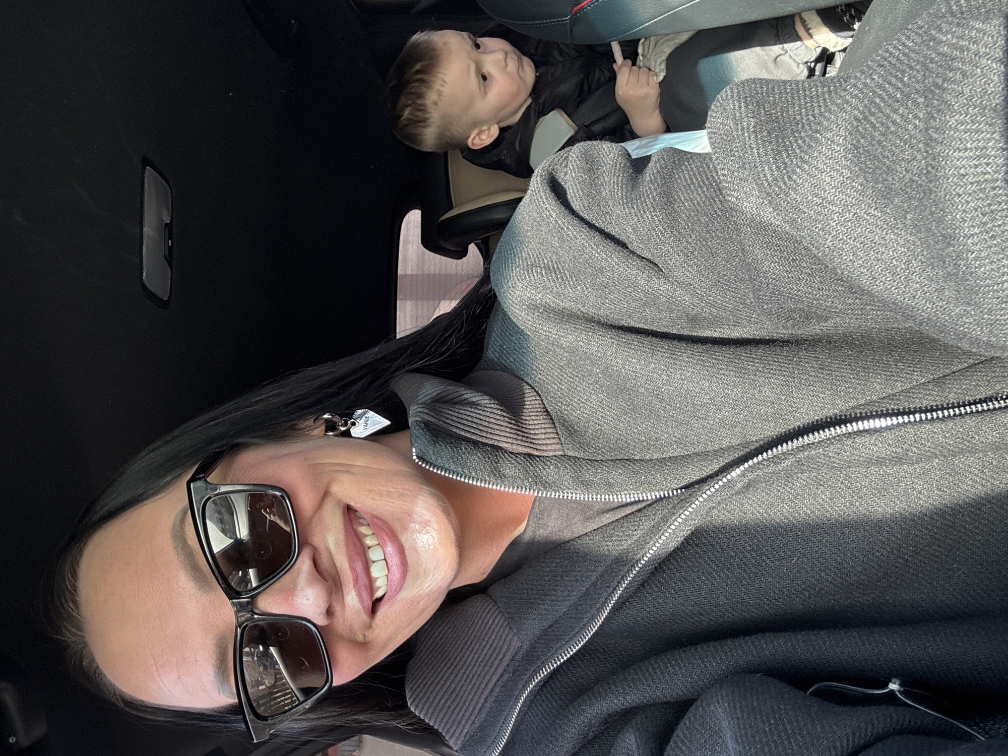The image displays the interior of a vehicle, likely a car, during daylight conditions. In the foreground, an adult female is positioned towards the left side of the frame, looking directly at the camera with a smile. She wears black framed sunglasses, a dark gray zippered jacket with a herringbone texture and a ribbed collar, and a silver heart-shaped earring inscribed with "VAHUE" on her right ear. Her dark hair is visible. In the background, to the right and partially obscured by the adult, a young male child is seated in a child safety seat. The child wears a dark jacket and holds a light-colored object in his right hand, gazing towards the right side of the frame. The vehicle's dark headliner and an overhead light fixture are visible. No specific location within the city of Chaikovsky, Russia, is discernible from the visual content; the scene is confined to the vehicle's interior.