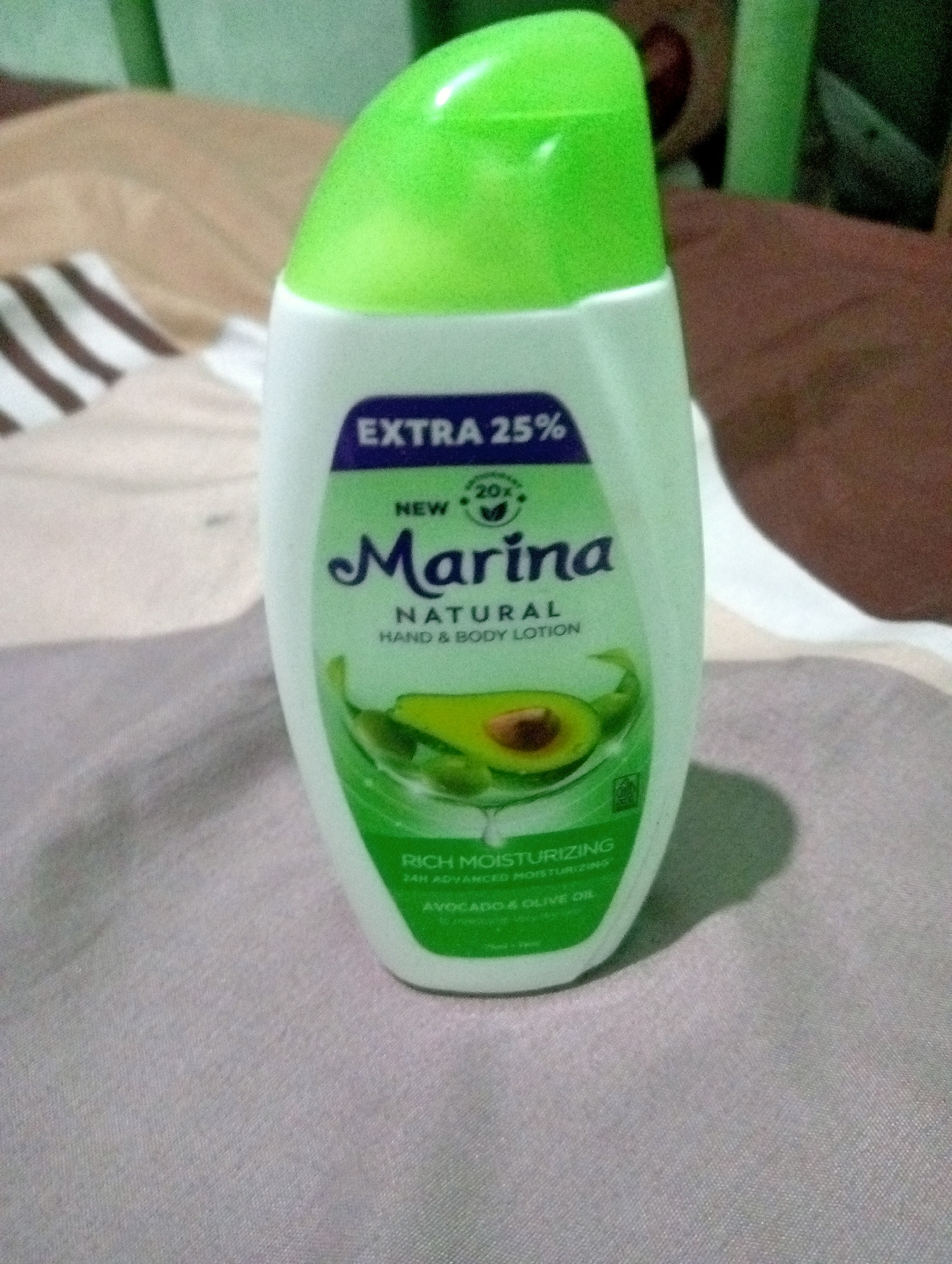 A white plastic bottle of Marina Natural Hand & Body Lotion with a green cap is placed on a striped fabric surface. The bottle has a label with the brand name "Marina" in dark blue lettering, and text indicating "EXTRA 25%", "NEW", "NATURAL", "HAND & BODY LOTION", "RICH MOISTURIZING", "24H ADVANCED MOISTURIZING", and "AVOCADO & OLIVE OIL". An image of sliced avocado is also visible on the label. The background is blurred, showing a combination of light brown and green elements. The scene is indoors, with no specific indicators of the city of Kota Negara Timur, Indonesia.
