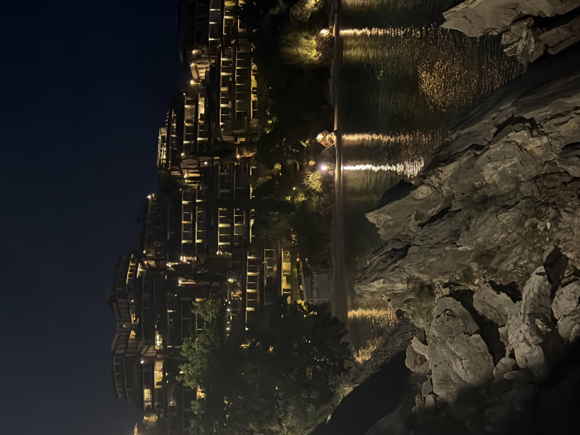A serene, vertically oriented nighttime photograph captures a luxurious coastal or lakeside resort in Budva, Montenegro. On the left side of the frame, a large, modern building complex with multiple terraced levels and balconies is brightly illuminated. Numerous warm, yellowish lights glow from within its windows and along its exterior, casting a vibrant light against the deep indigo sky. The architecture suggests a resort or high-end residential property, thoughtfully integrated into the natural landscape.
To the right of the illuminated buildings, a calm body of water, likely the sea or a large bay, stretches across the middle ground. The still surface perfectly reflects the architectural lights, creating striking vertical streaks and shimmering patterns of light across the dark water. Scattered trees and foliage, some subtly lit, are visible along the waterfront and between the buildings.
In the foreground and extending along the right edge of the water, rugged, light-colored rock formations are prominent. These natural, jagged rocks add a dramatic and organic element to the scene, contrasting with the man-made structures. The overall impression is one of peaceful nocturnal beauty, showcasing a sophisticated development nestled within a picturesque natural setting. The clear, dark sky and the bright artificial illumination indicate that the photograph was taken at night. No people or visible text are present in the image.