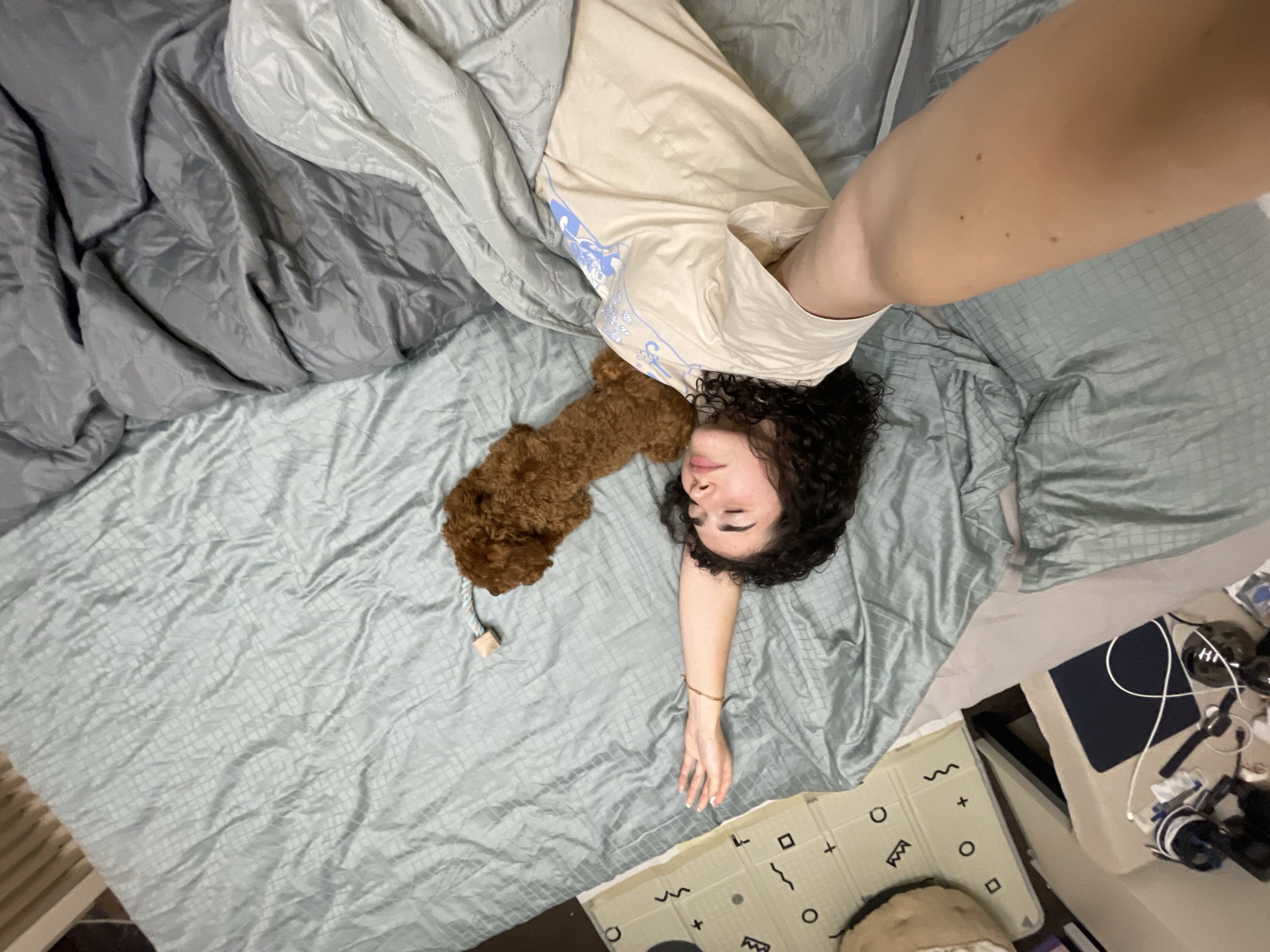 A top-down view captures a person lying on a bed. The bed is covered with a pale blue-green patterned bedsheet. The person, presumably female, has dark curly hair and is wearing a beige t-shirt. A brown poodle dog is lying near her head. A gray comforter is partially covering the person on the left side of the frame. The person's arm extends downwards along the bed.
To the right of the bed, on the floor, is a patterned playmat and a bedside table with various objects, including a laptop, cables, and other miscellaneous items.
There is no information visible in the picture to suggest that the image was taken in Sofia, Bulgaria.