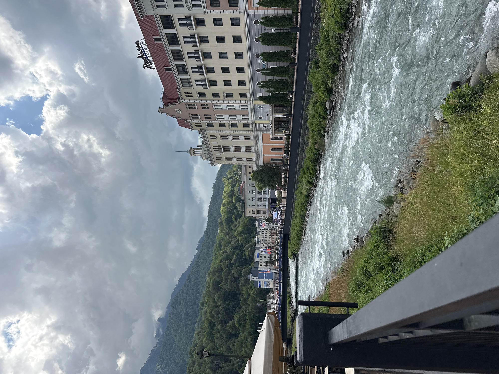 The image depicts a wide, fast-flowing river in Estosadok, Russia, with a modern hotel on the right bank and mountains covered in lush green trees in the background. The hotel, identified by a "Radisson" sign, is a multi-story building with a red roof and a light-colored facade. The river's turbulent water suggests a strong current. On the left side of the image, a dark railing and a table with a beige umbrella indicate an outdoor seating area, likely a cafe or restaurant. A bridge spans the river in the mid-ground, connecting the two sides of the town. The sky is overcast with dramatic clouds. Several cypress-like trees are planted in a row along the riverbank in front of the hotel. People can be seen walking along the promenade beside the river. The overall scene suggests a scenic resort town, possibly in a mountainous region.