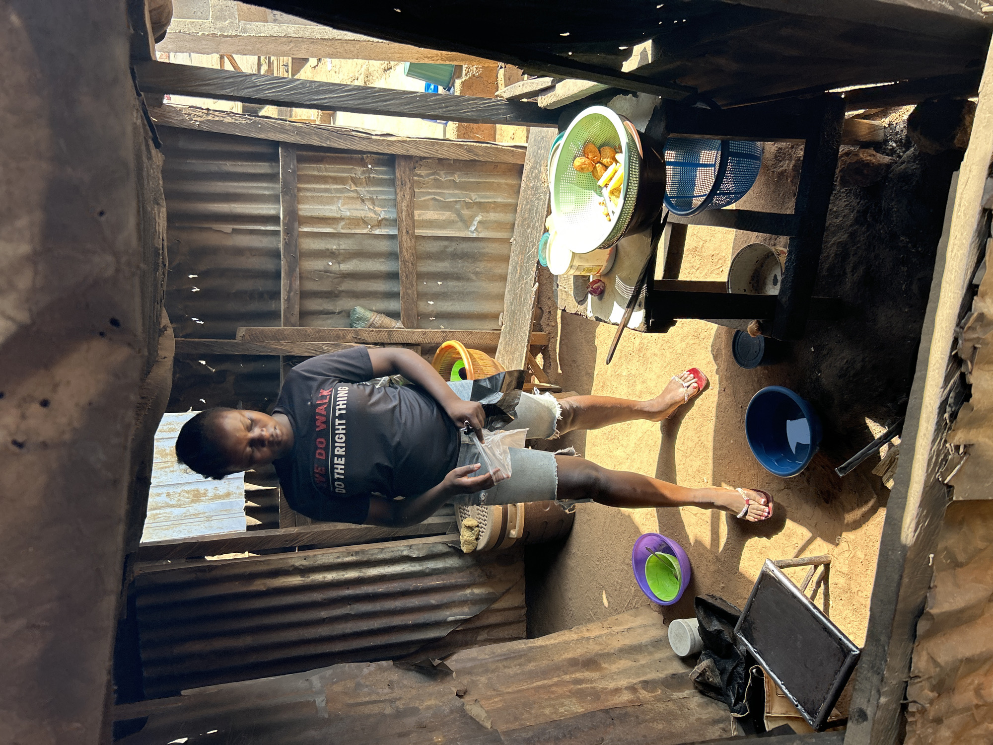 The image shows a person standing in what appears to be an outdoor kitchen or food preparation area in Jos, Nigeria. The structure has walls made of corrugated metal sheets. The person is wearing a dark t-shirt with white lettering that says "WE DO WALK DO THE RIGHT THING," denim shorts, and red sandals. They are holding a clear plastic bag in their hands. The kitchen space contains several bowls of varying colors, a basket with what looks like food items, and a cooking stove. The area seems cluttered and basic. The lighting is uneven, with strong sunlight entering from above and casting shadows.